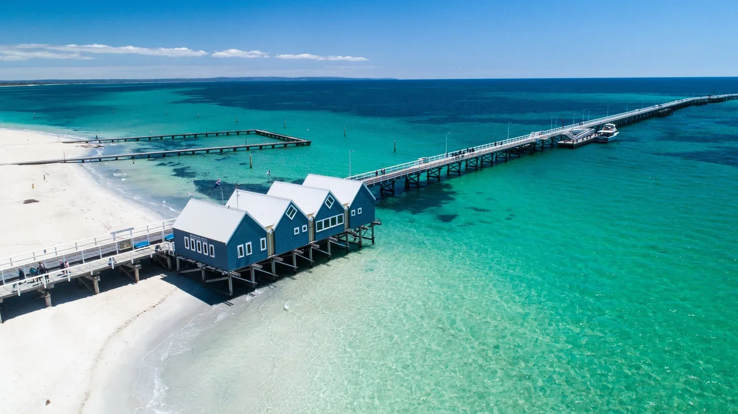 An aerial view of a pier with blue beach houses extending into a turquoise sea, with a sandy beach nearby and a clear blue sky overhead.