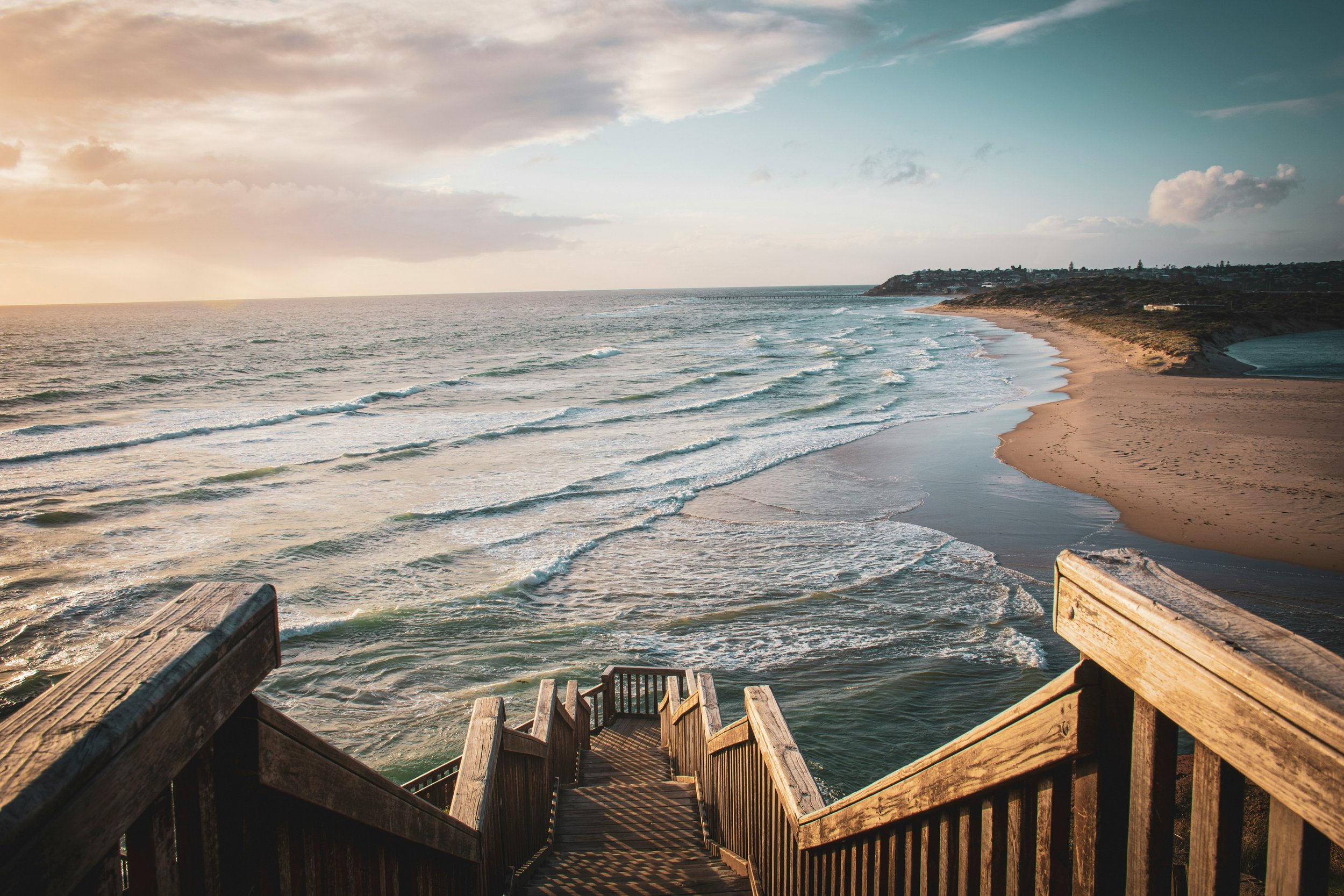 View of a beach from a wooden staircase overlooking the shoreline with waves and a distant coastal hill.