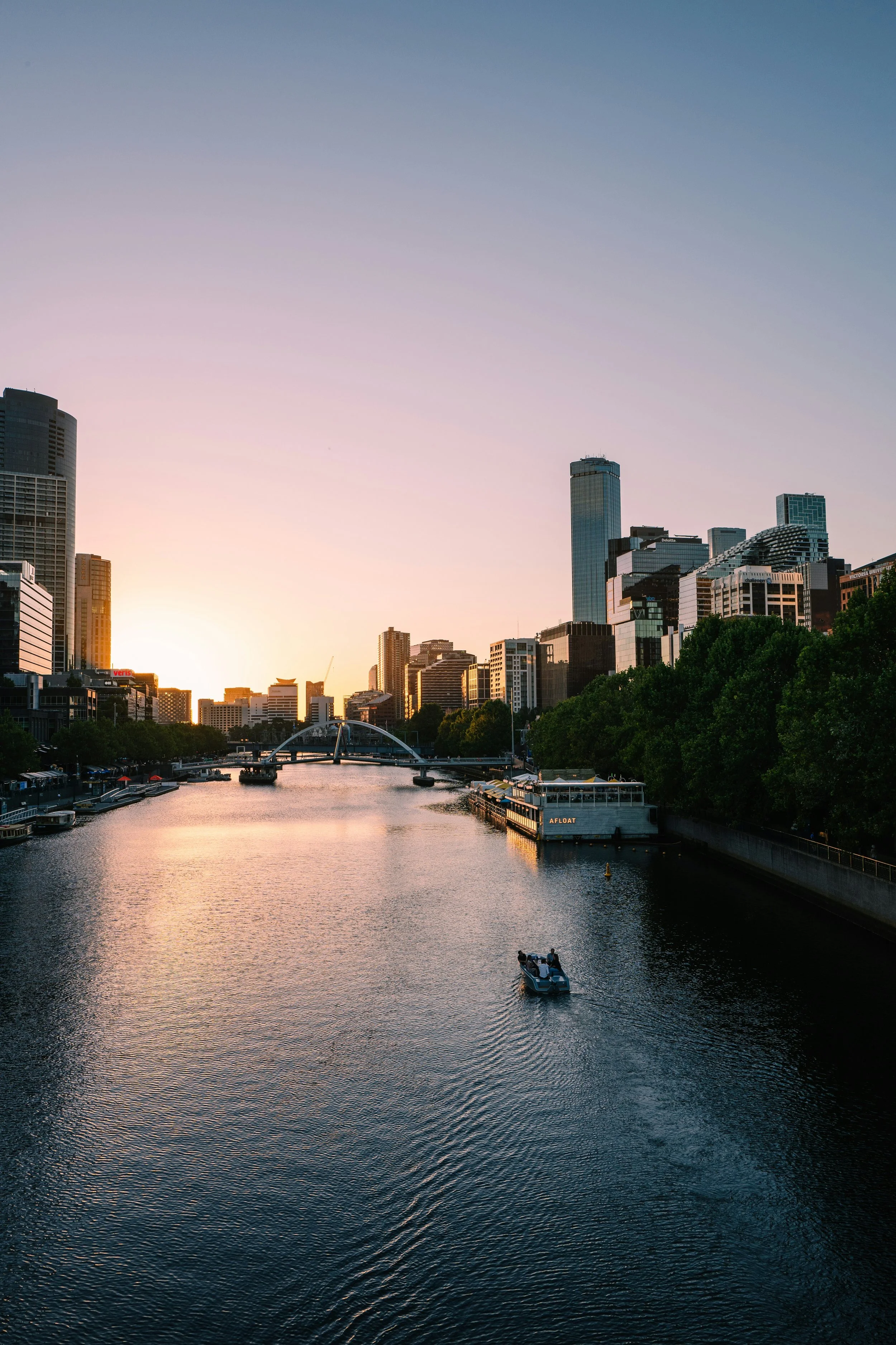 City skyline at sunset with tall buildings, a river with a boat, and a bridge in the foreground.