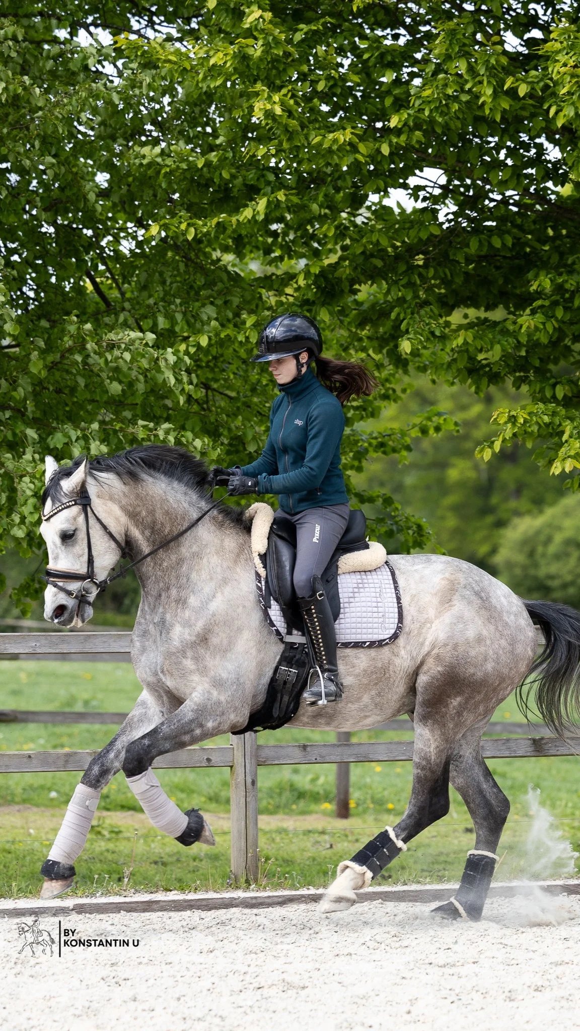 Ein Mädchen beim Reiten auf einem grauen Pferd im Grünen, mit Helm und Reitausrüstung, in einer Reitbahn auf holprigem Boden.