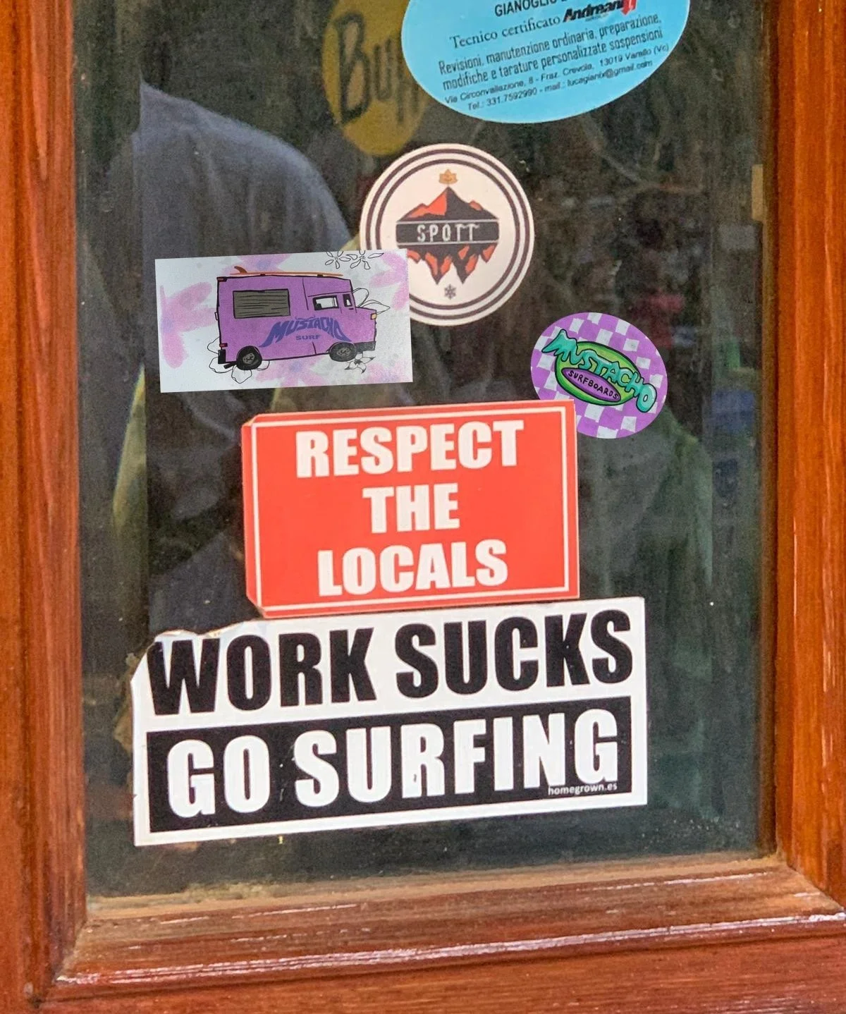 Fenster mit Aufklebern: 'Respektiere die Einheimischen', 'Arbeit ist Mist, geh surfen', und Surfmarken