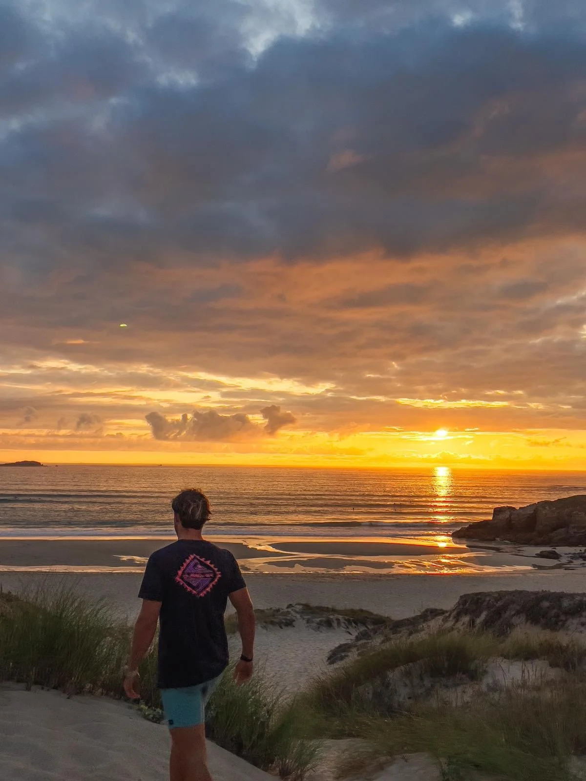 Mann schaut auf einen Sonnenuntergang am Strand mit Wolken am Himmel und einer spiegelnden Wasserfläche im Hintergrund.