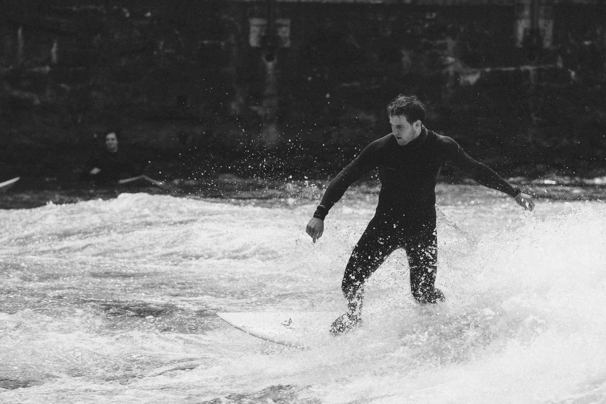 Ein junger Mann in einem Neoprenanzug steht im Wasser und schaukelt auf einem Surfbrett, während Wellen gegen das Brett schlagen. Im Hintergrund ist eine Person im Wasser zu sehen.