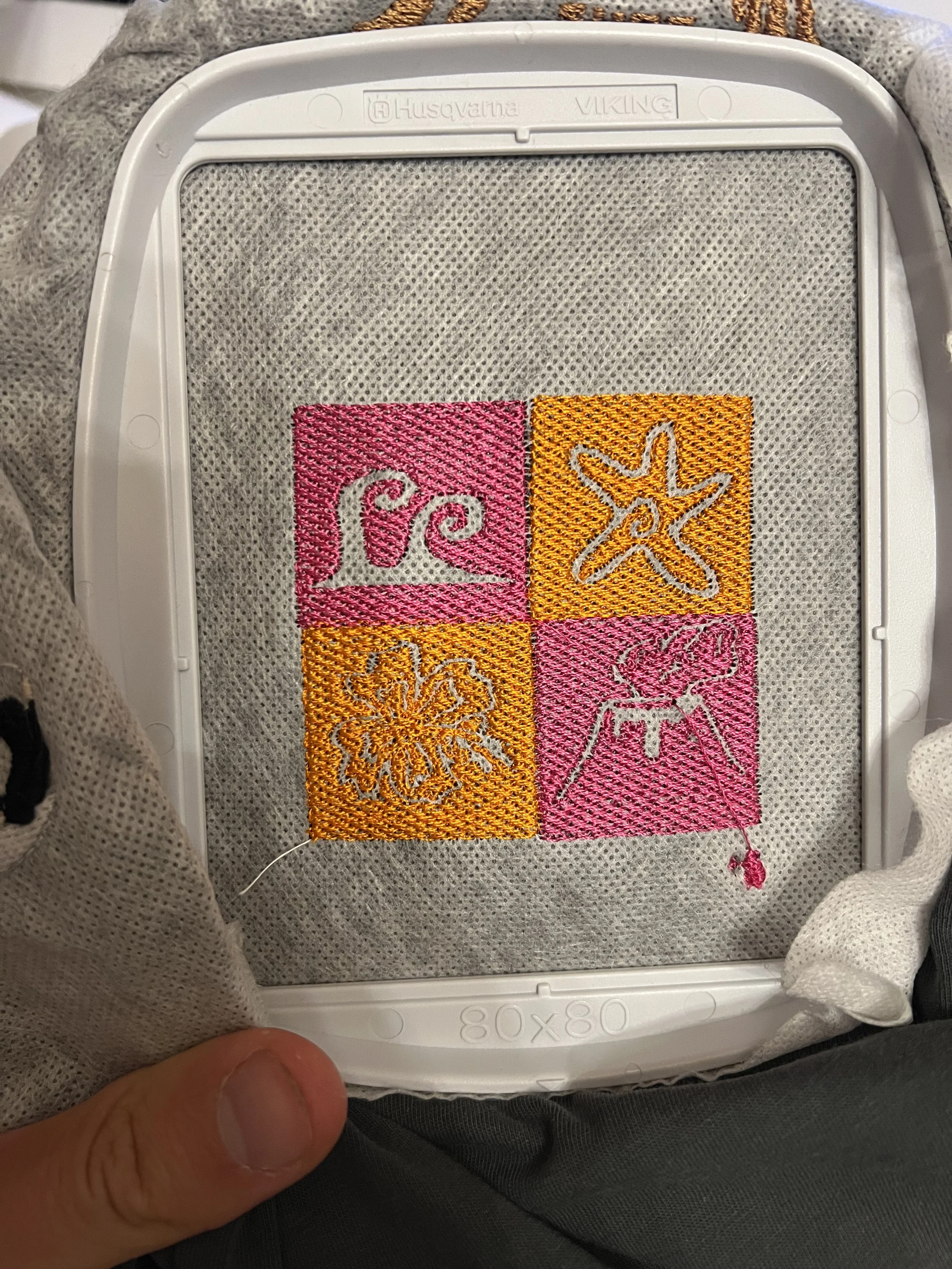 Hand, die ein gesticktes Stoffstück mit vier Quadranten hält, die jeweils ein Muster und eine prominente Figur enthalten, auf einer weißen Kunststoffbox. Der Stoff ist in Pink und Orange gehalten.