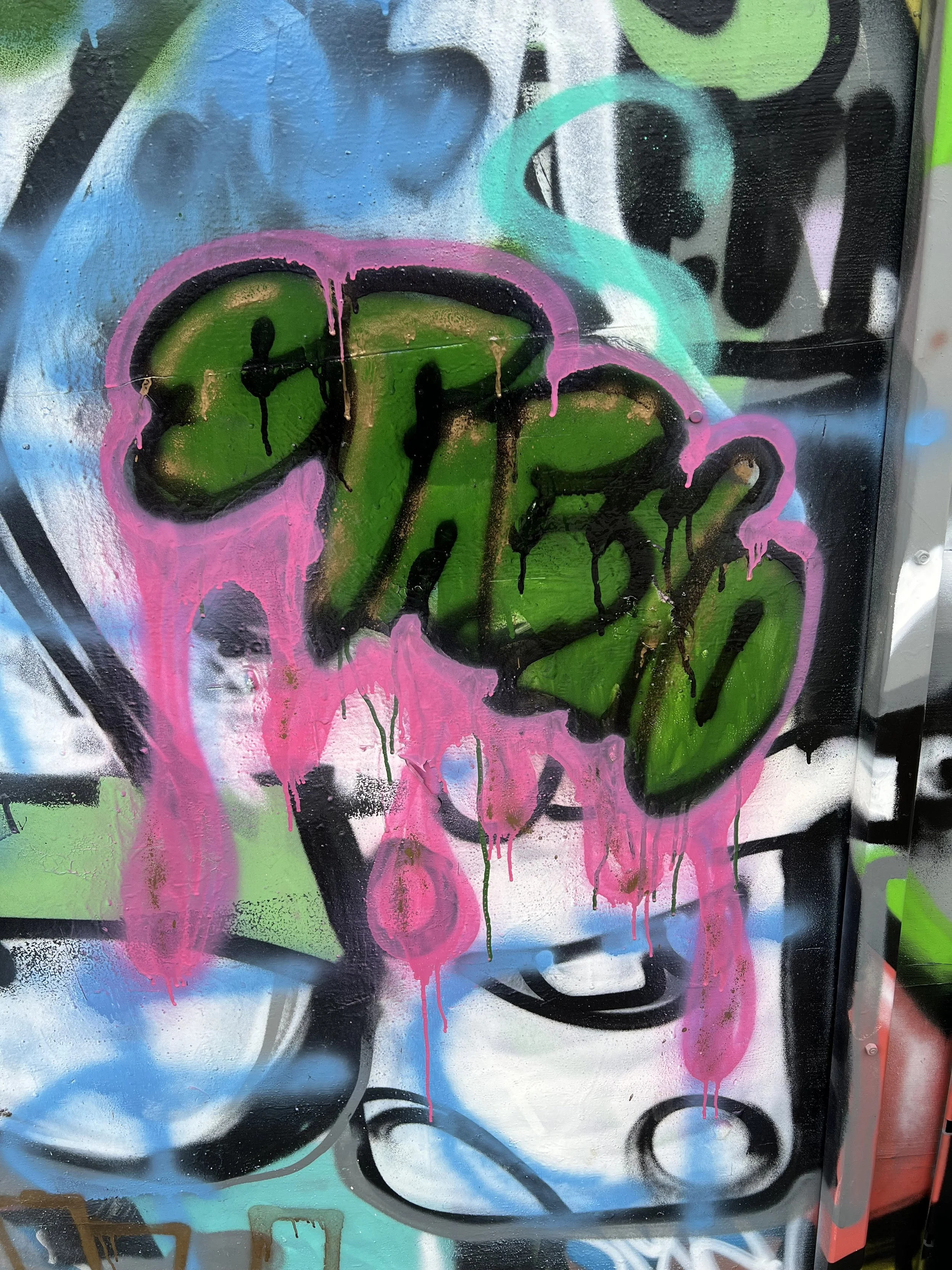 Graffiti mit einem stilisierten, pink umrandeten Logo, umgeben von bunten Farben und dripping Effekten auf einer Wand.