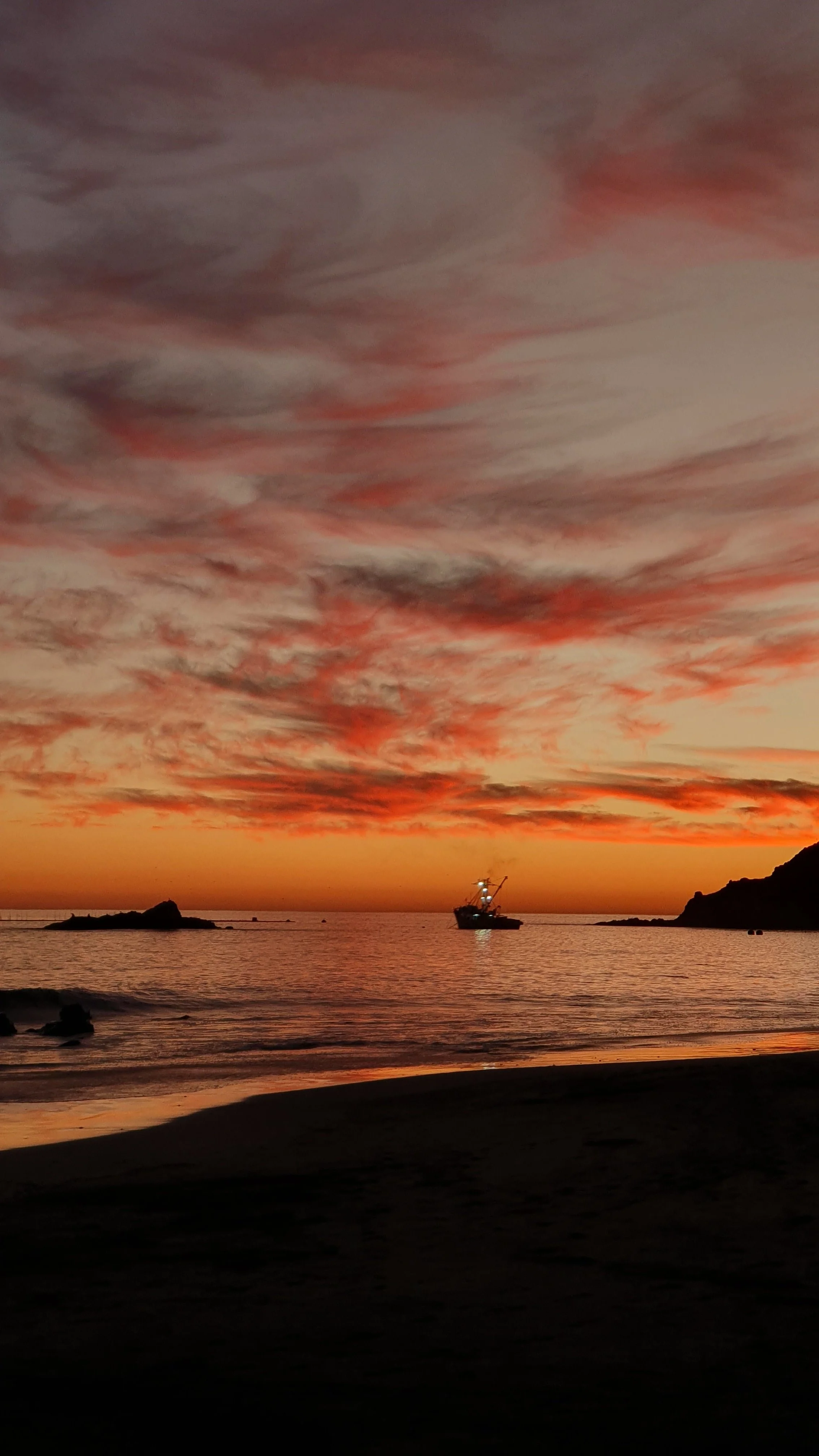 Eine Küstenlandschaft bei Sonnenuntergang mit einem Schiff im Wasser, dunklen Felsen und Wolken am Himmel in Orange, Rot und Grau.