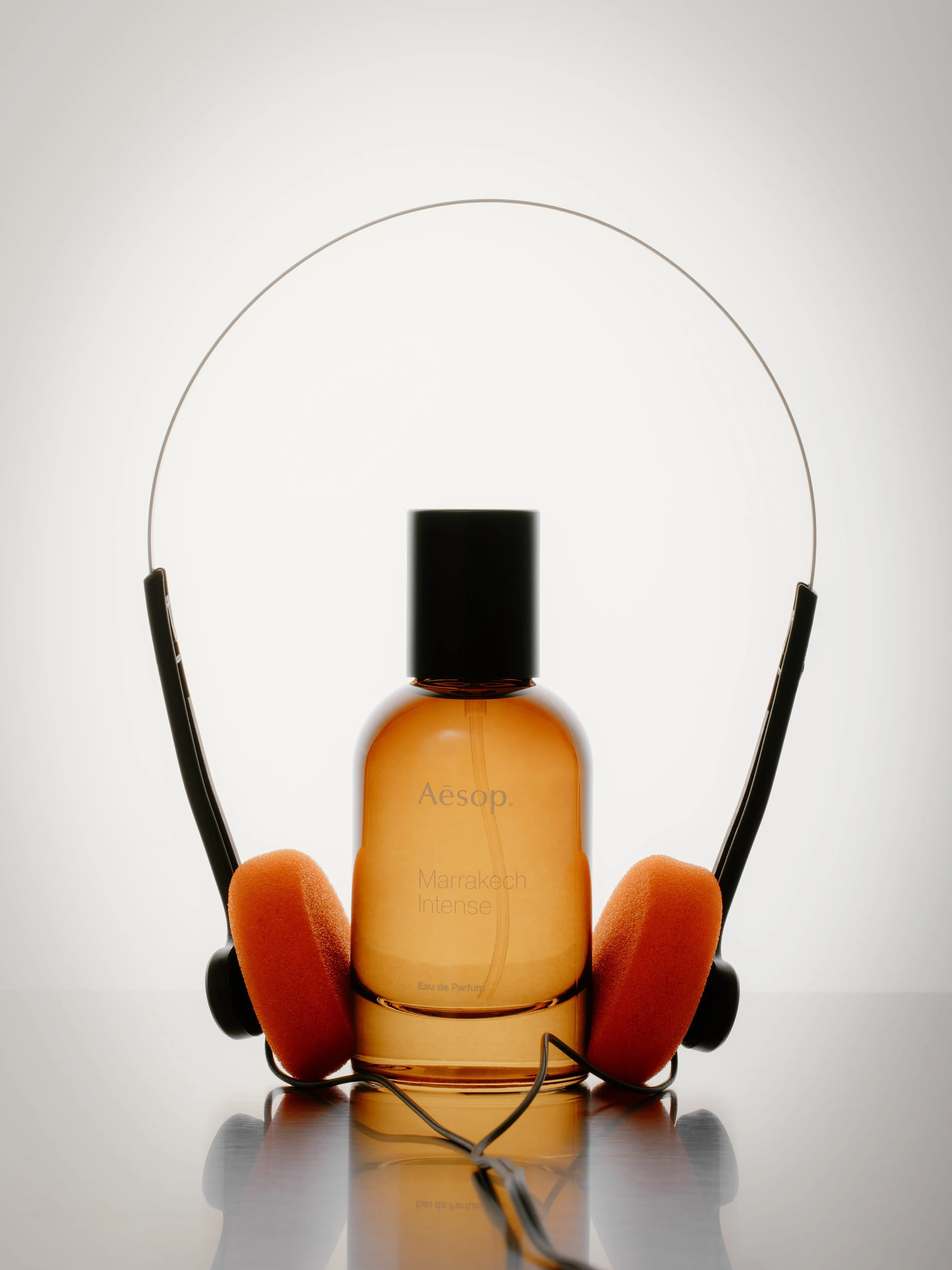 A bottle of Aesop Marrakech Intense Eau de Parfum is held by a hand wearing orange gloves, with a pair of black headphones surrounding the bottle against a plain white background.