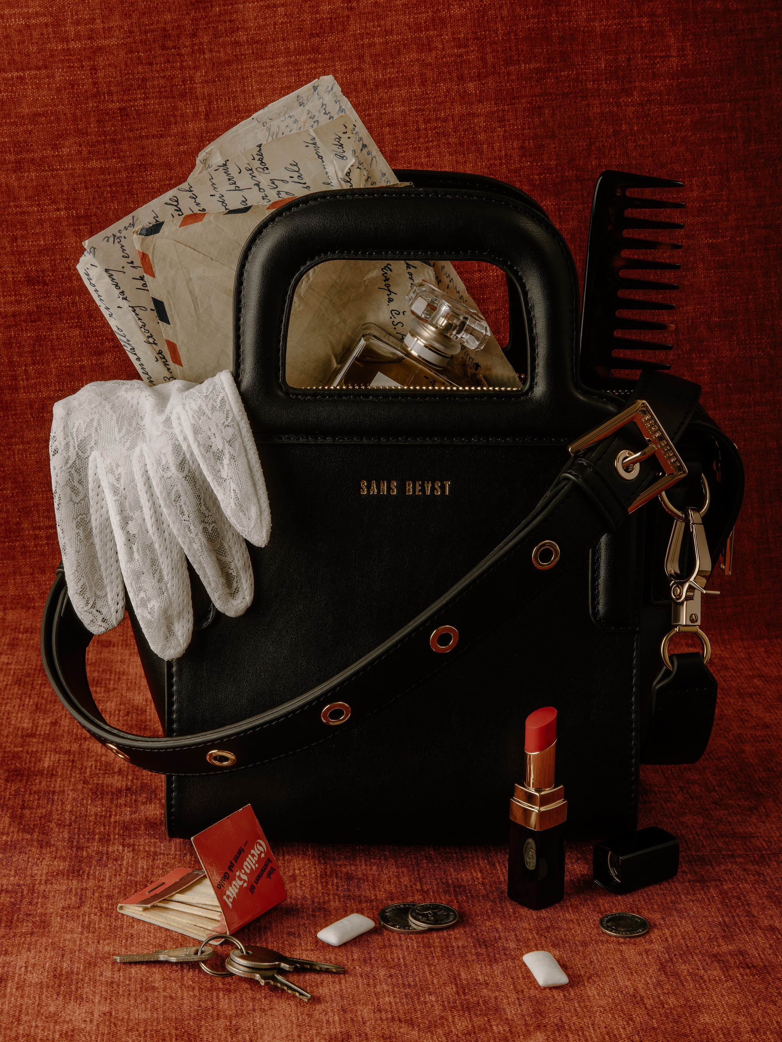 A black handbag with the words SANS BEVST on it. Inside the bag, there is a small glass perfume bottle, a piece of paper with handwritten text, and a black hair comb. Outside the bag, on a red textured surface, there are white lace gloves, a red lipstick, a small white eraser, some coins, a set of keys, a lipstick and a black object.