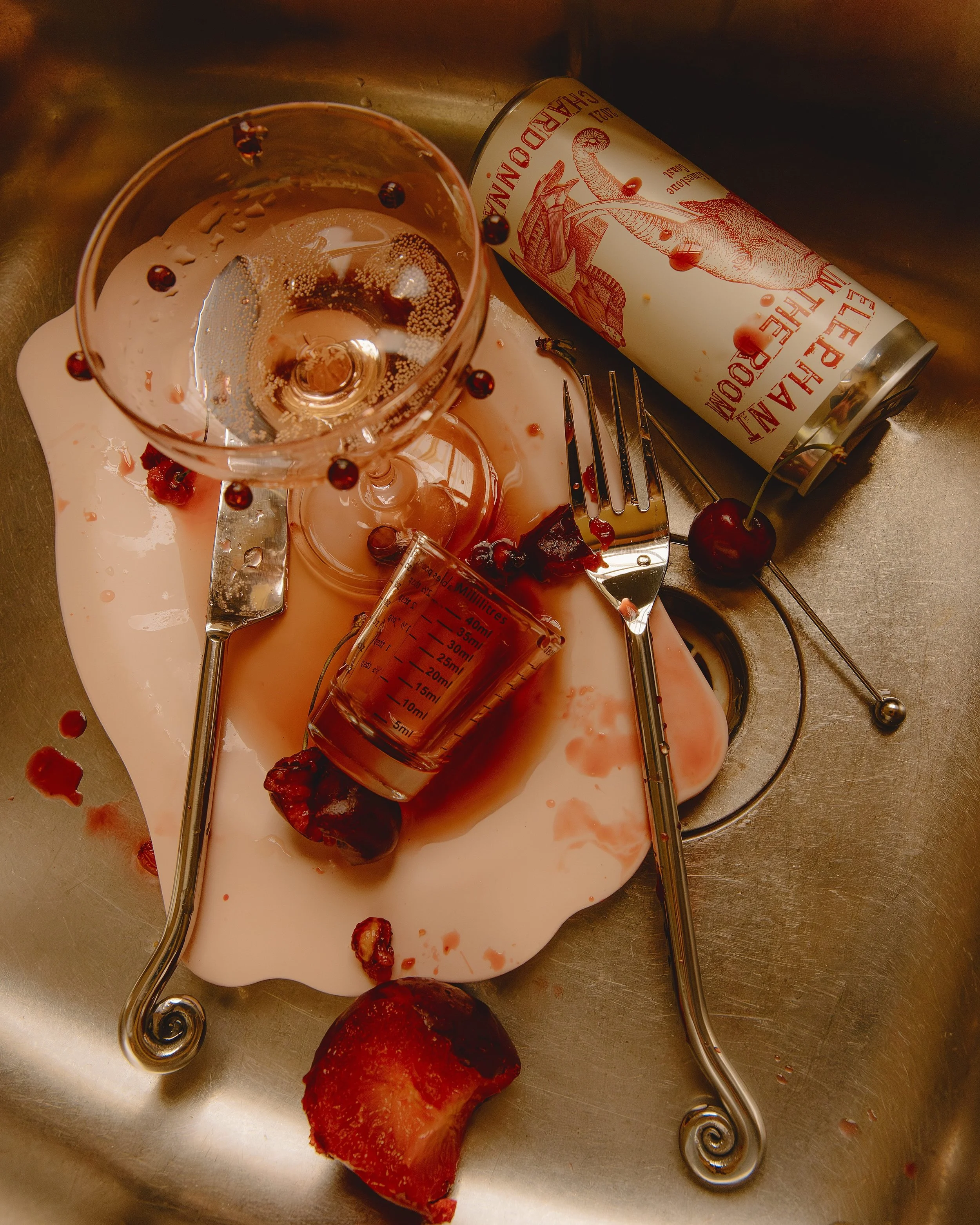 A spilled glass of rosé wine, a can labeled "Leham's Hard Drink" with a dinosaur illustration, a glass measuring cup, a fork, and some cherries scattered around in a metallic sink.