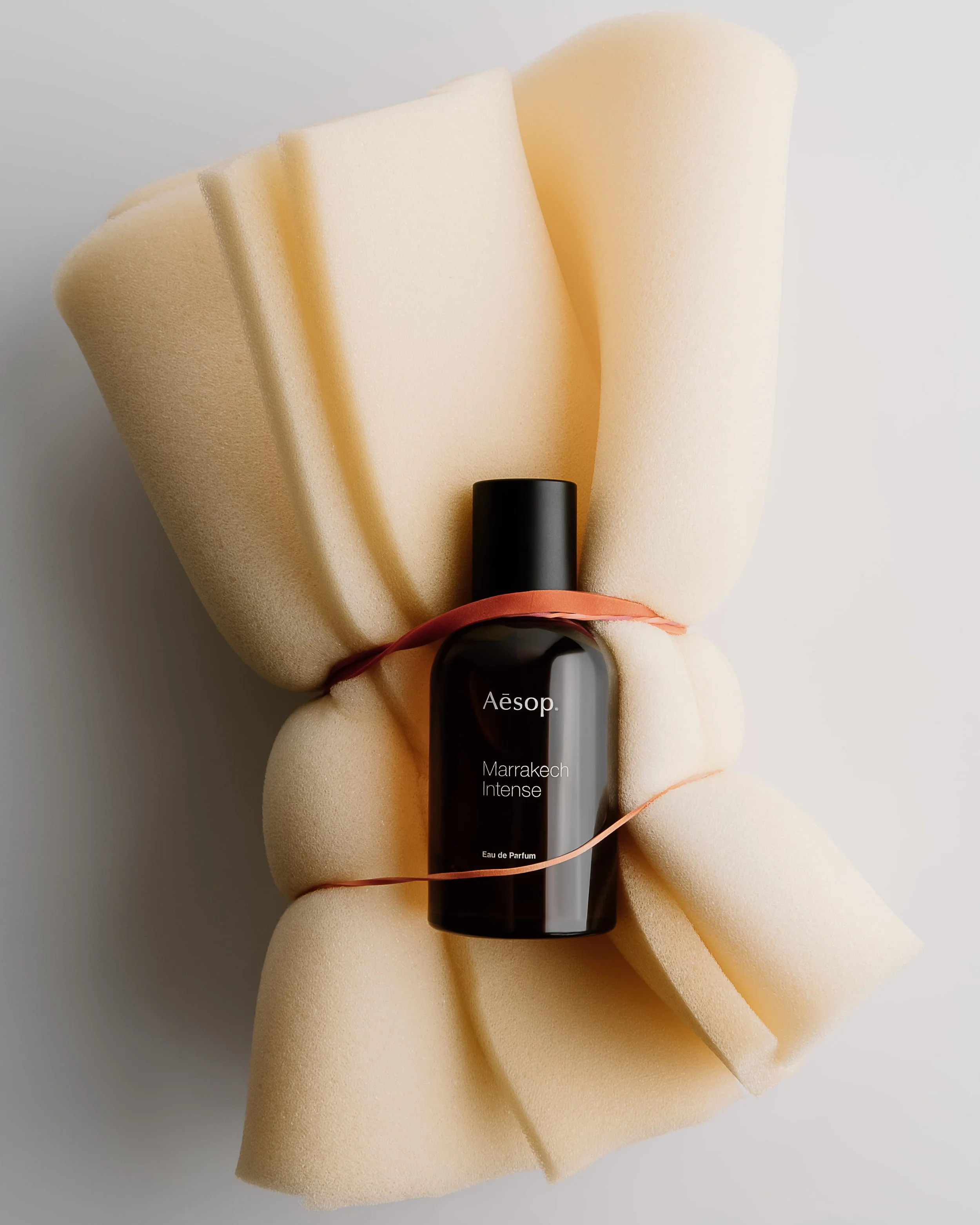 A hand holding a black bottle of Aesop Marrakech Intense Eau de Parfum, surrounded by foam sheets and a pink rubber band, against a white background.