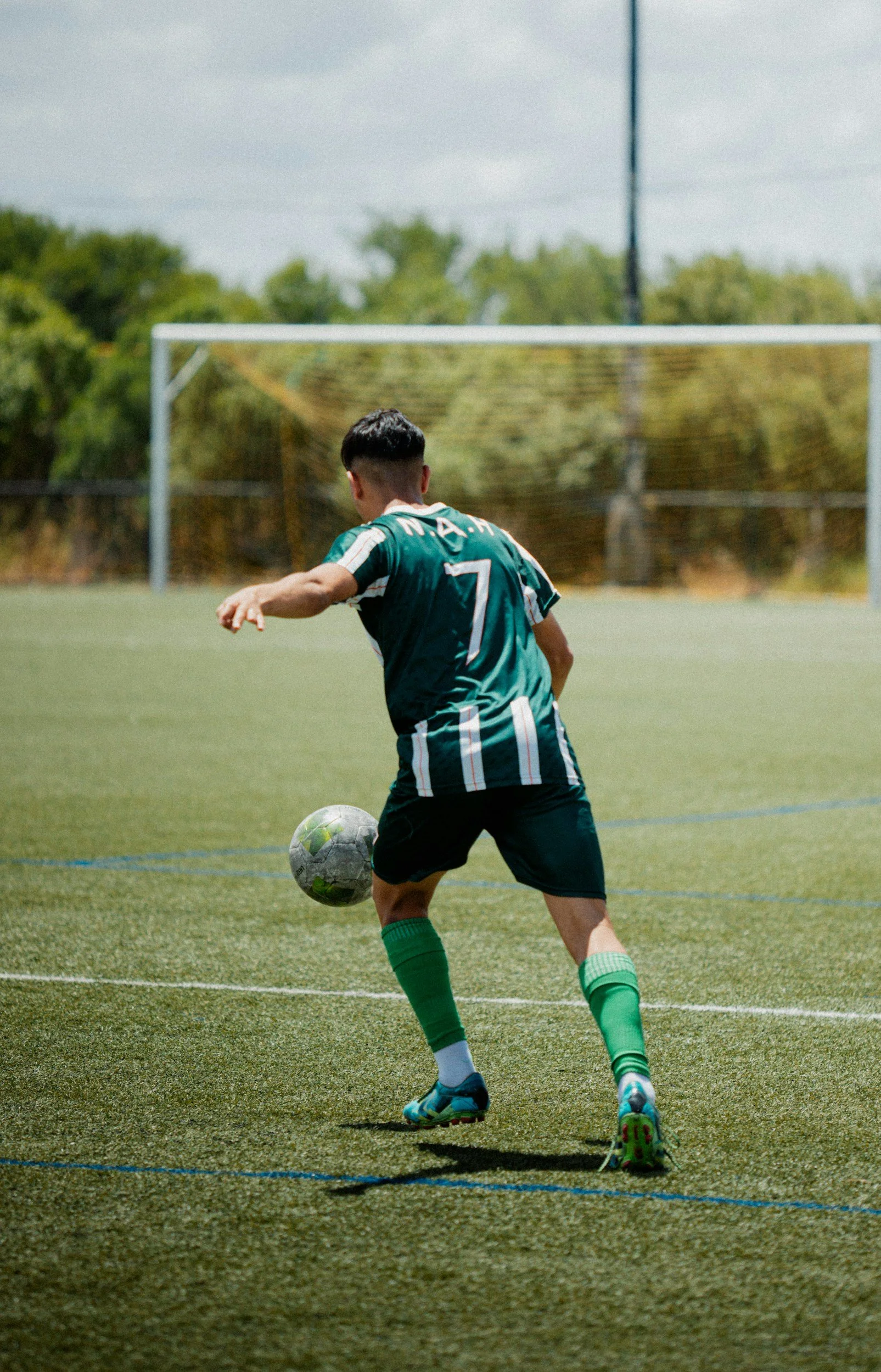 A soccer player in a green uniform with the number 7 on his back is kicking a soccer ball on a field with a goal in the background.
