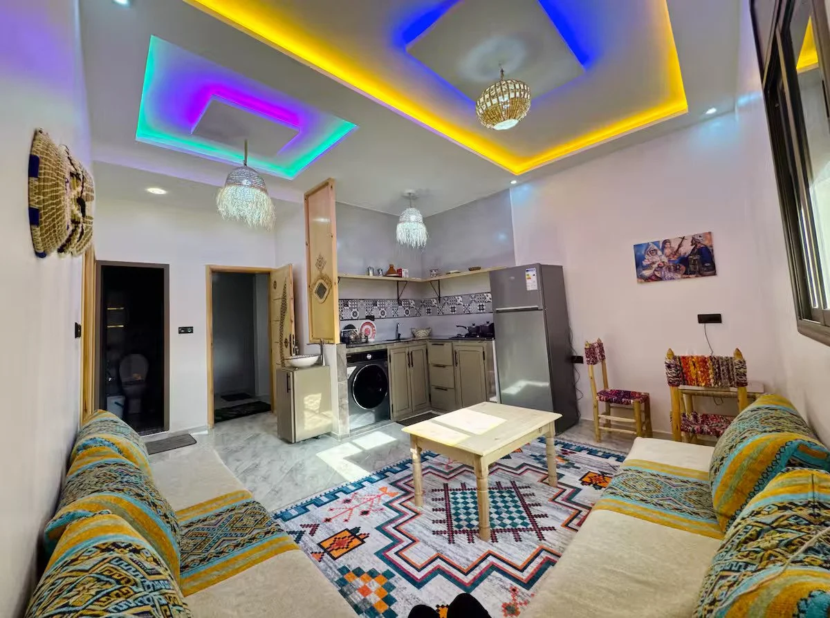 Living room with sofas, a coffee table, patterned rug, kitchen area with gray cabinets, refrigerator, and hanging lamps. Colorful LED ceiling lights and a window on the right.