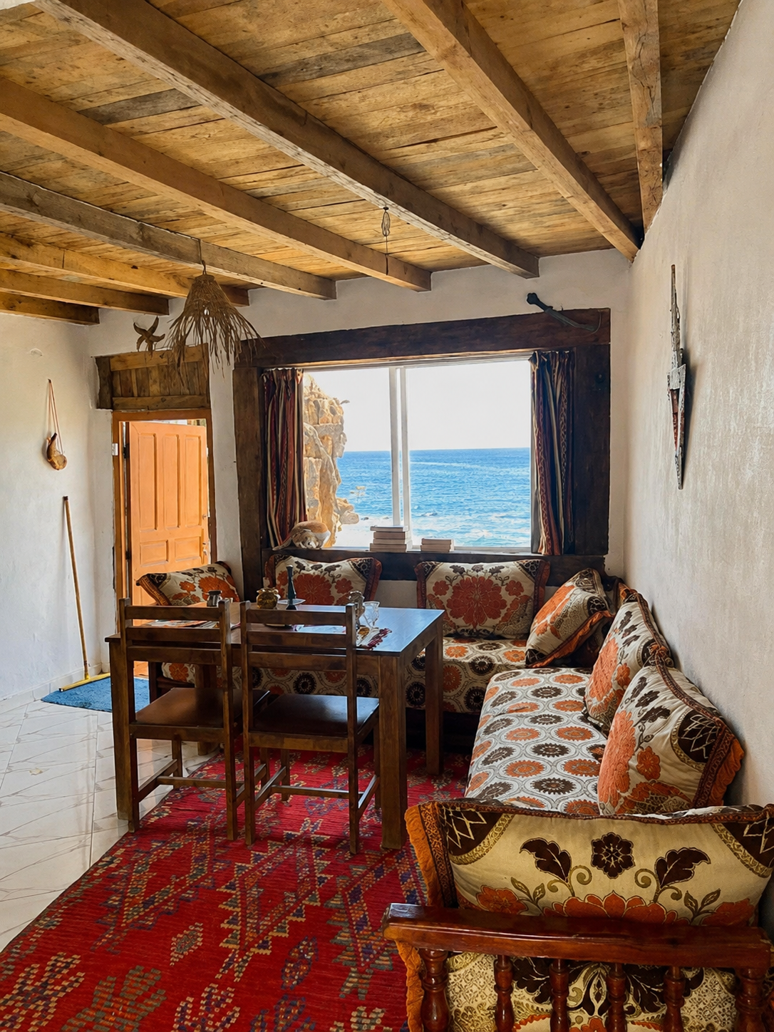 Cozy living room with a view of the ocean through a large window, featuring wooden ceiling beams, patterned sofas, a wooden dining table with chairs, and a red rug.