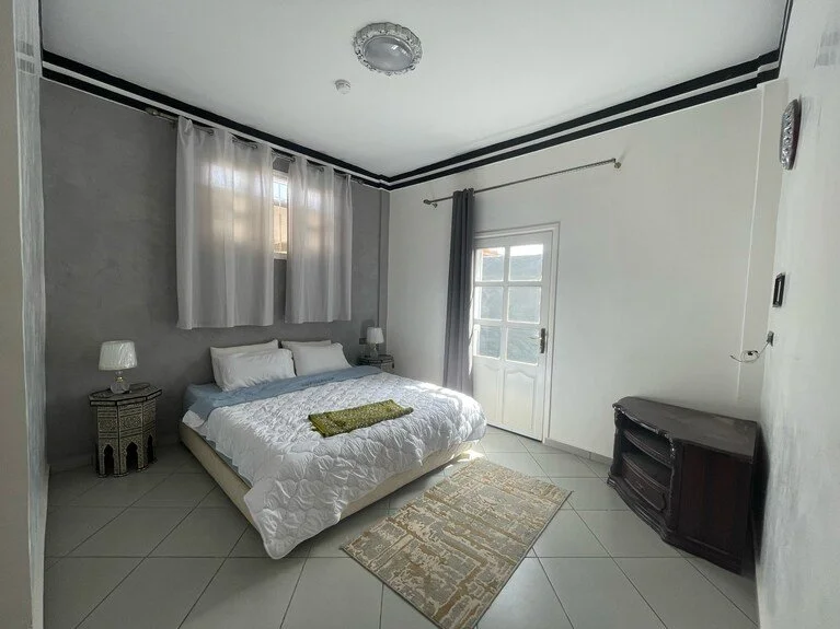 A bedroom with a white bed, gray nightstands, white walls with black trim, two windows with curtains, a door, a small rug, and a television stand.