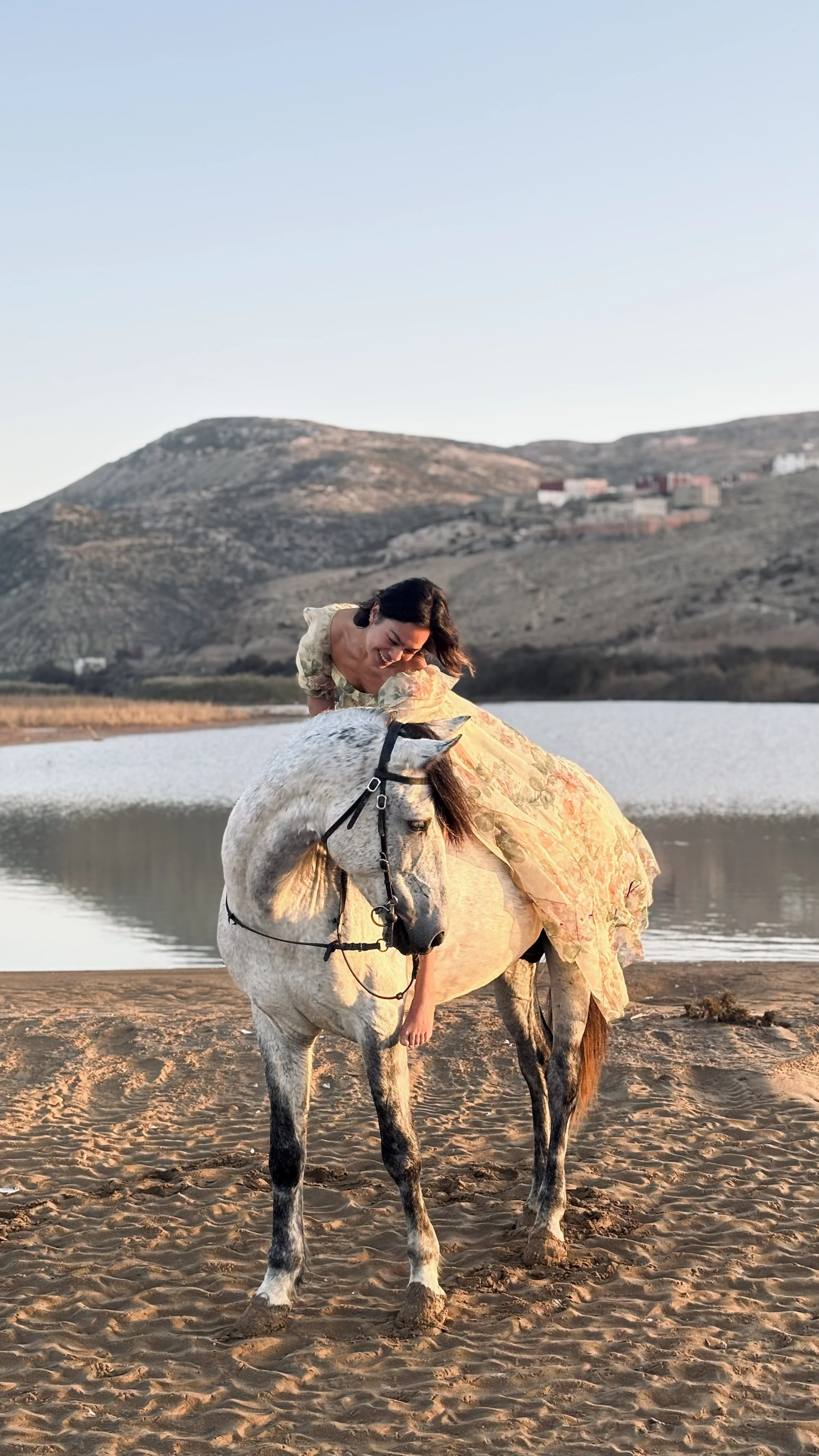 A woman in a floral dress riding a white horse on the sandy shore of a lake with mountains and houses in the background during sunset.