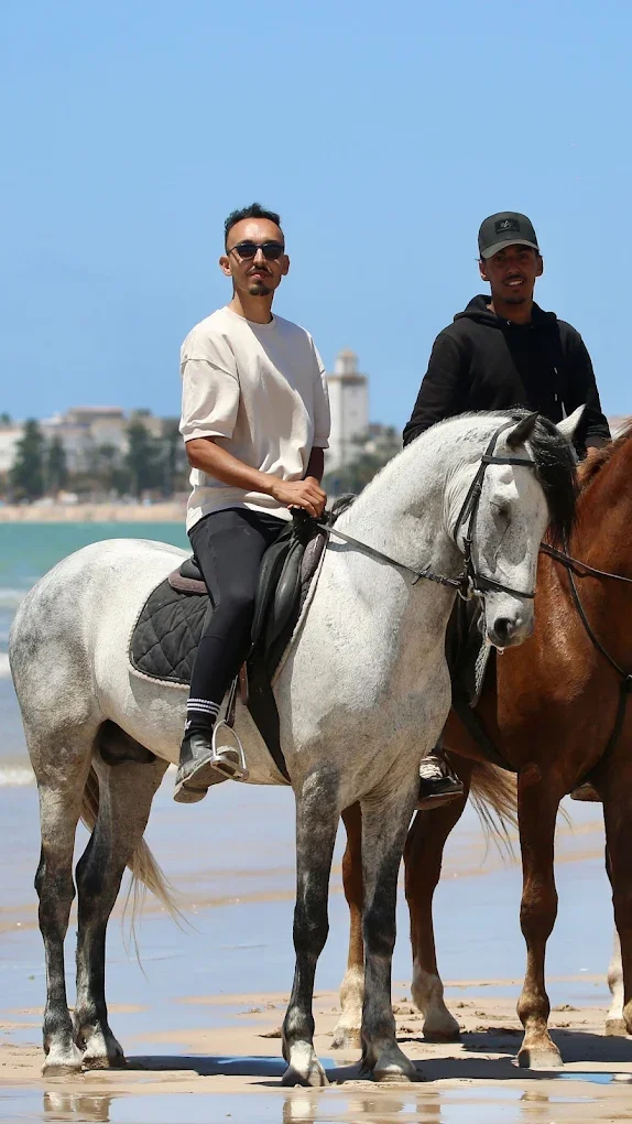 Two men riding horses on a beach with water and buildings in the background.