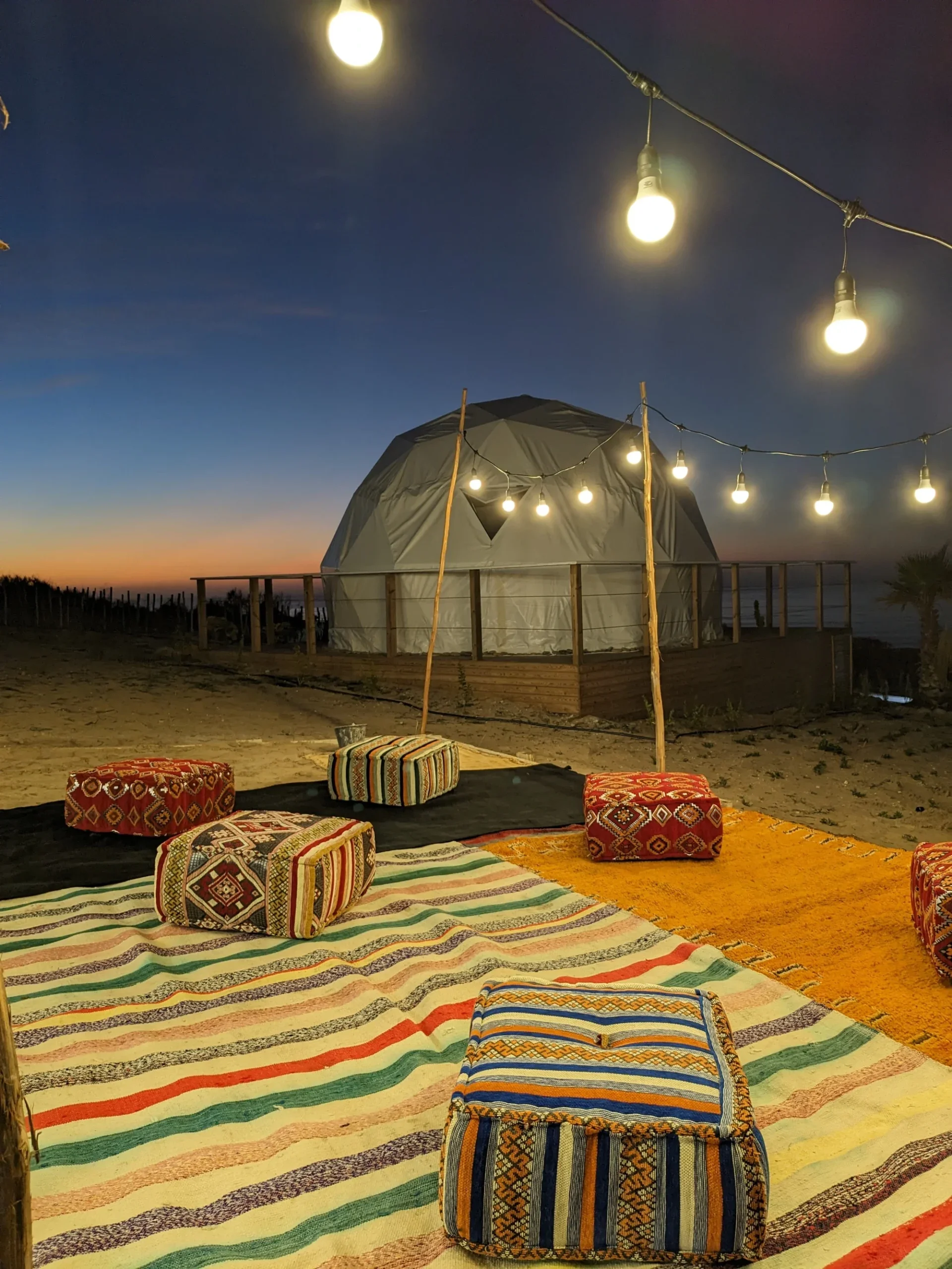 An outdoor scene at dusk or night with string lights hanging above a wooden deck or platform. On the ground, there are colorful patterned cushions and rugs. In the background, there is a small tent or yurt with an open door, illuminated from within. The sky is darkening, with a hint of sunset on the horizon, and a few trees or plants are visible near the horizon.