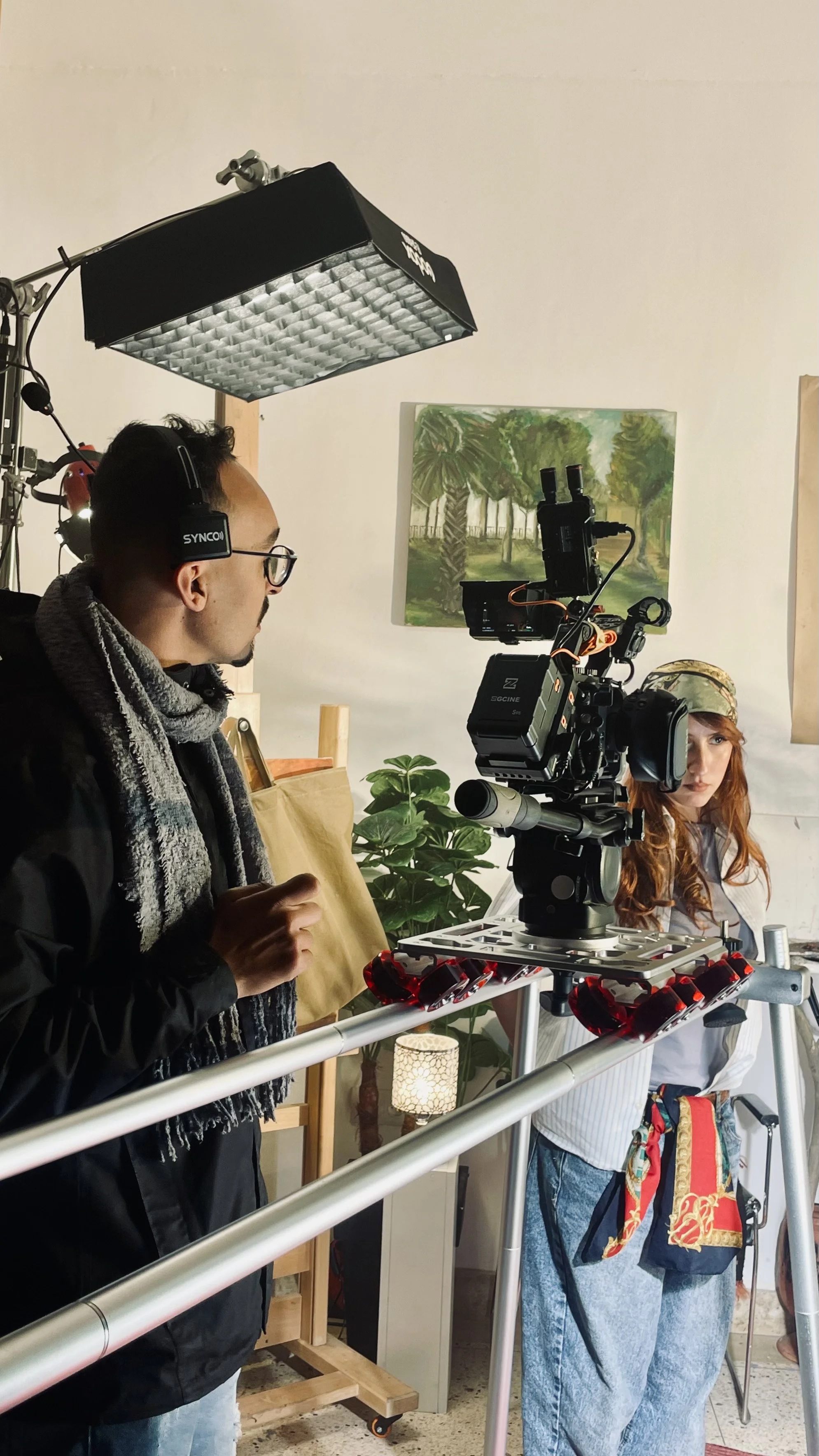 A film crew setting up equipment indoors. A man wearing glasses and headphones operates a professional camera on a tripod. A woman stands nearby, dressed in casual clothes with a colorful bag. The background includes a painting of a forest scene and a plant.