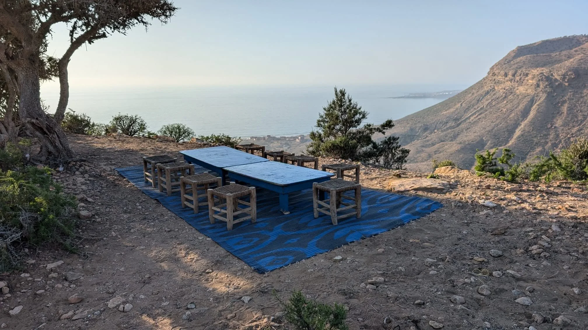 Outdoor seating area with a blue table and small wooden stools on a blue rug, overlooking a mountain and body of water in the distance.