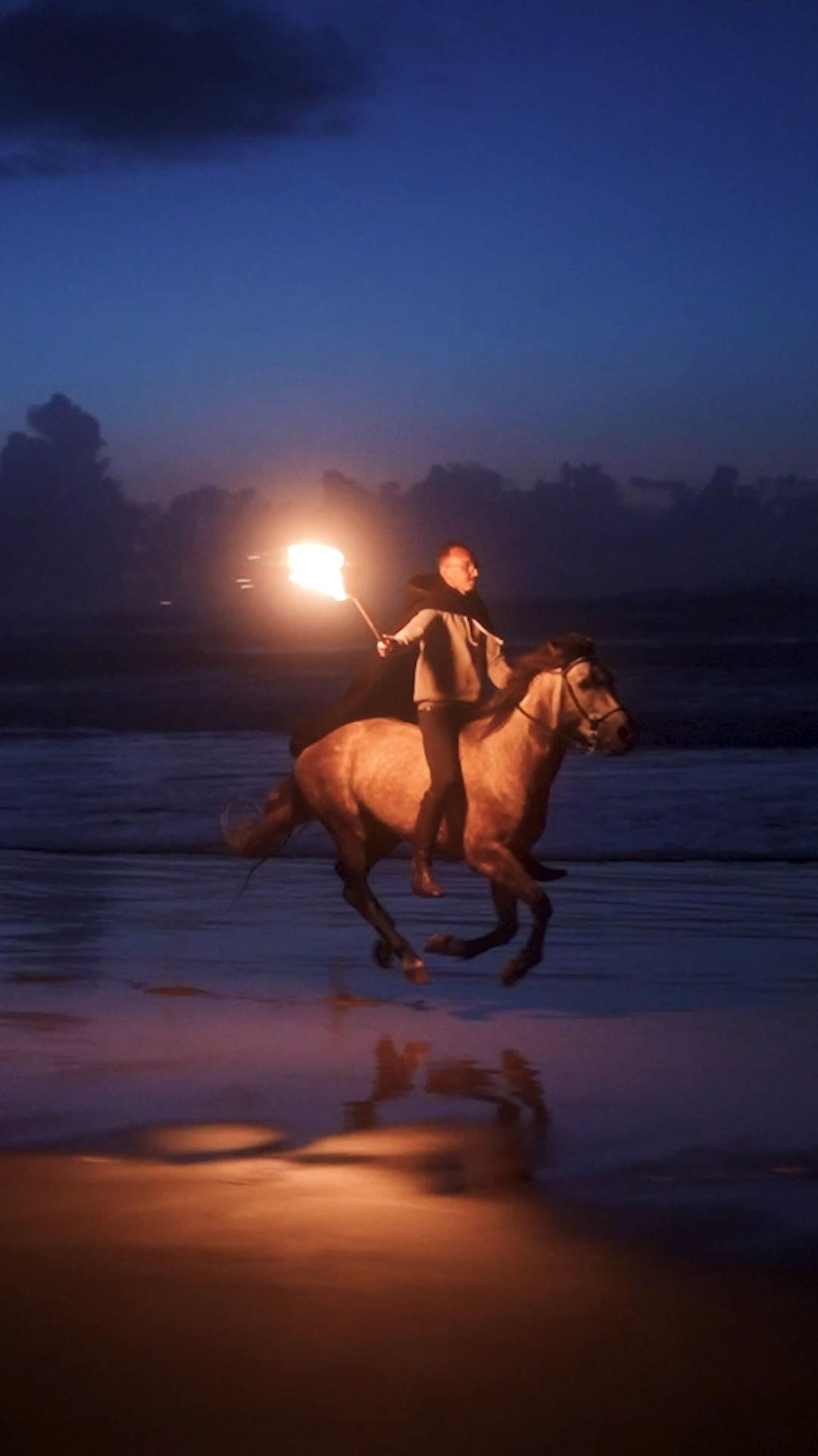 Person riding a horse on the beach at night holding a lit torch.