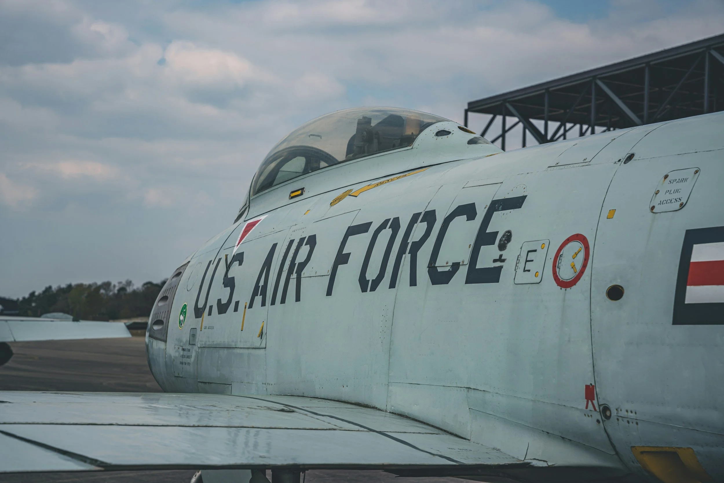 Ein Flugzeugrumpf mit US Air Force Beschriftung, im Hintergrund ein Himmel mit Wolken und eine Lagerhalle.