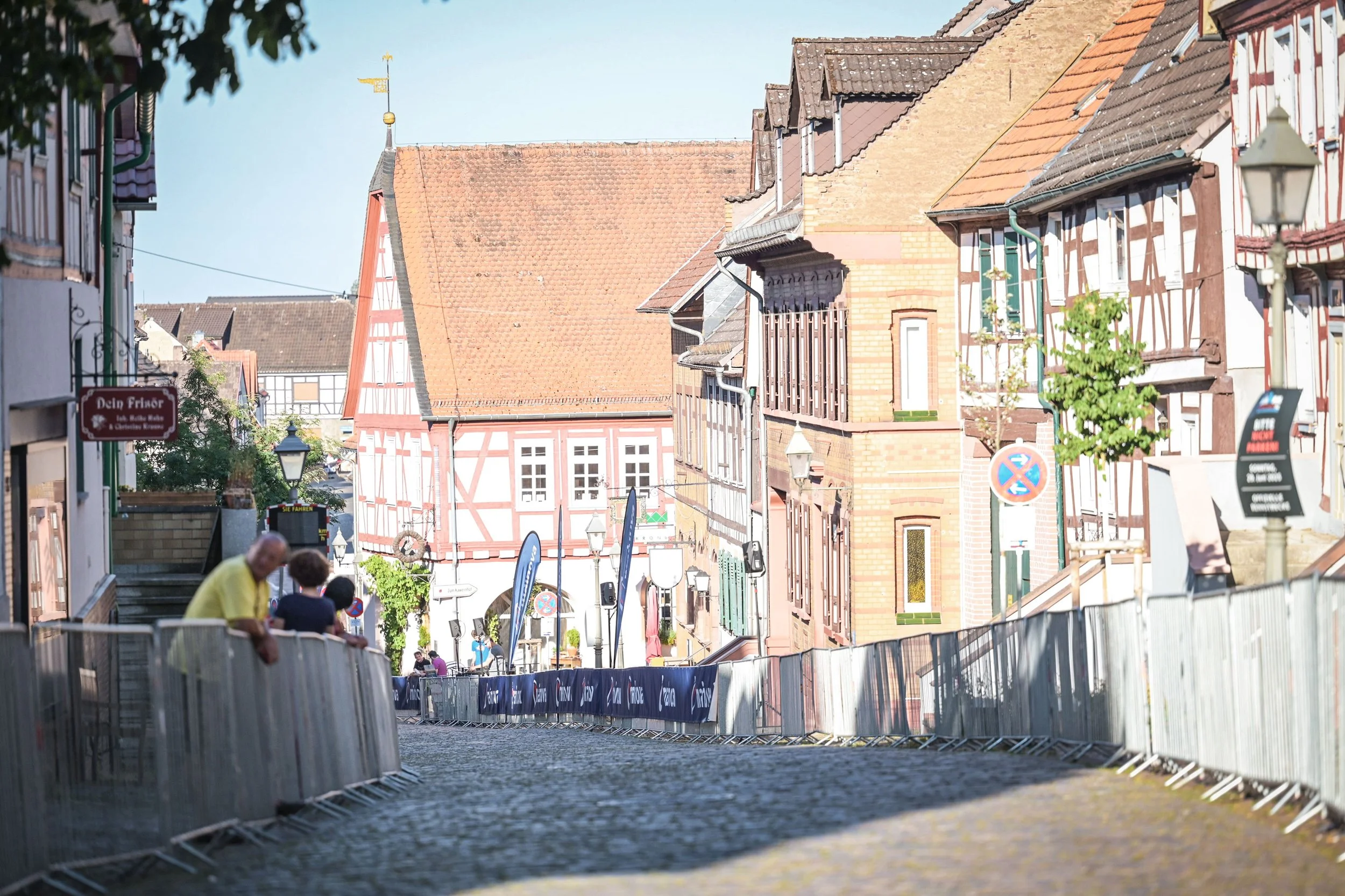Straße in einer historischen Stadt mit alten Fachwerkhäusern und roten Dächern, Menschen gehen entlang der Absperrungen, und es gibt Laternen und Geschäfte an der Seite.