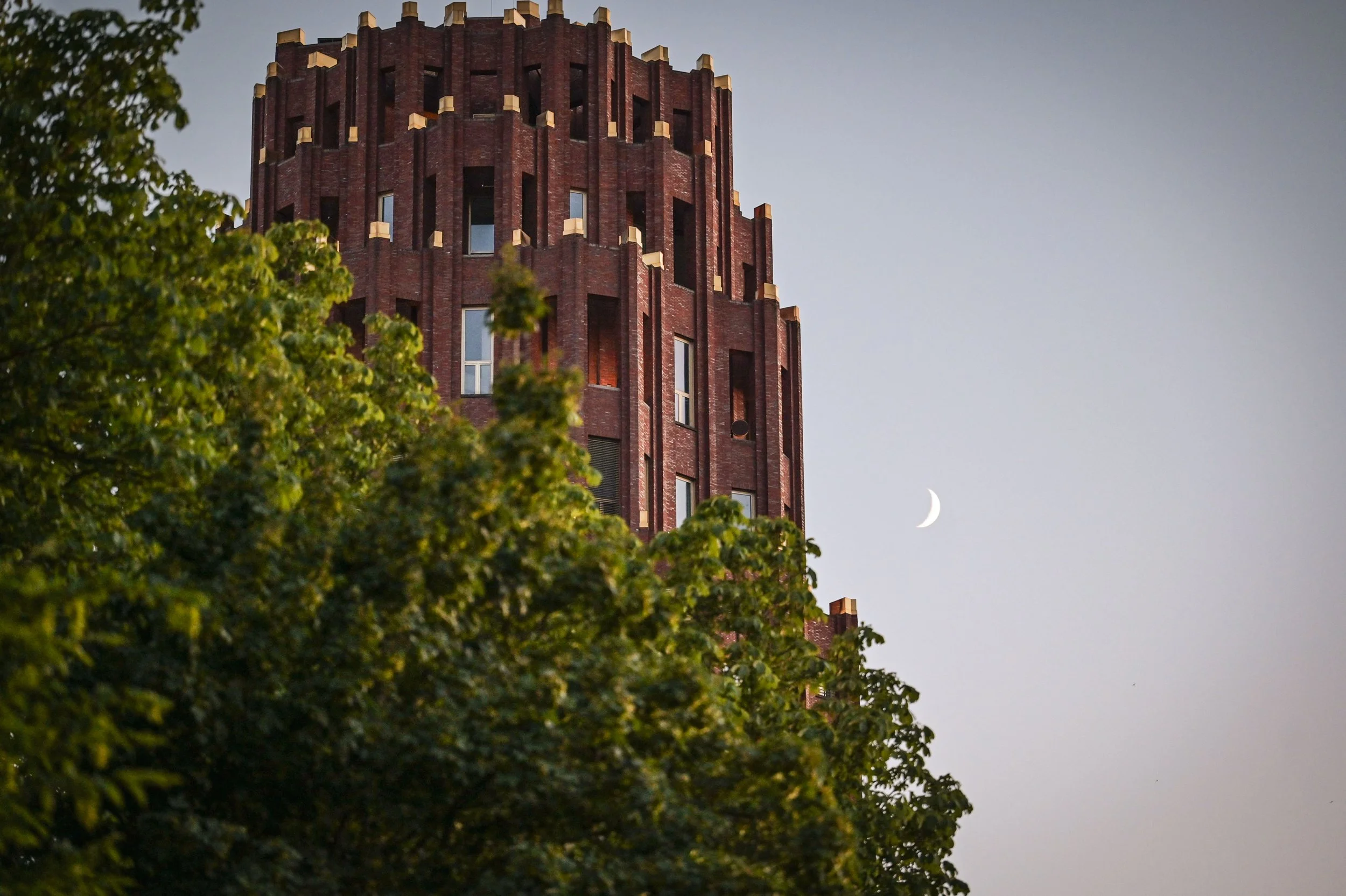 Ein rotes Backsteingebäude mit einer ungewöhnlichen runden Architektur, umgeben von grünen Bäumen, bei Dämmerung, mit einer sichtbaren Mondsichel am Himmel.