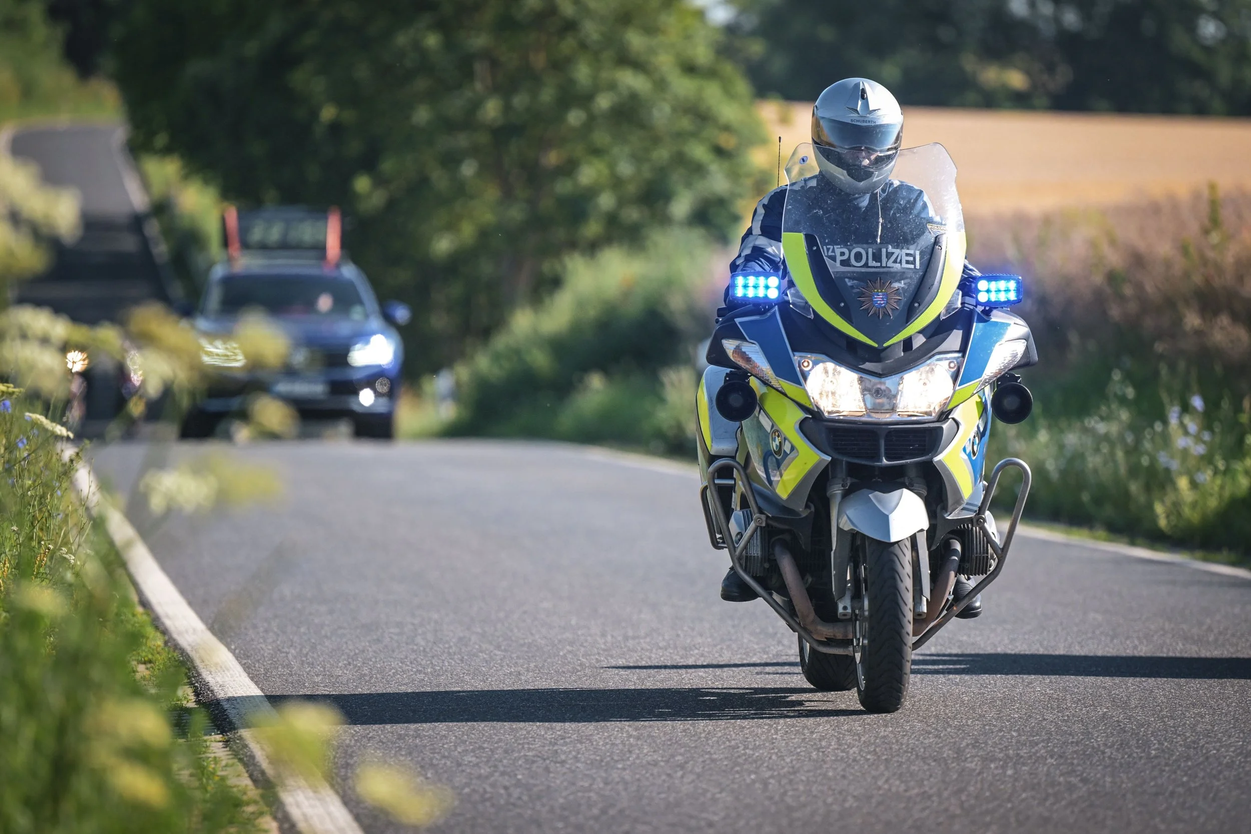 Polizeimotorrad auf einer Straße mit grünen Bäumen im Hintergrund, blauer Blaulicht leuchtet.