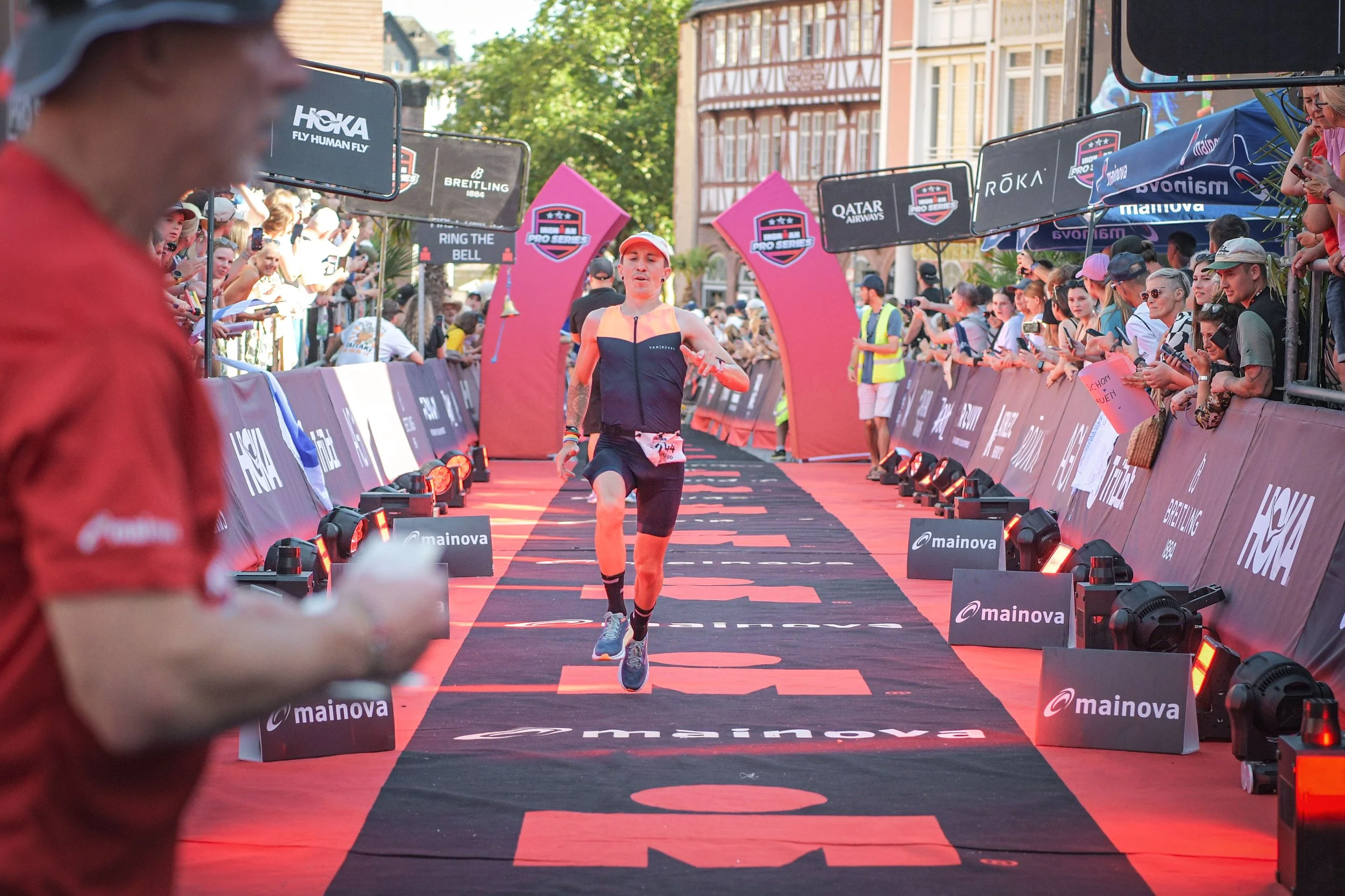 Ein männlicher Athlet läuft am Ziel im Rahmen eines Triathlons. Er trägt einen schwarzen Anzug mit Fan-Designs, eine Mütze und einen Zeitmesser am Handgelenk. Die Szene ist bei Sonnenuntergang mit einer Menschenmenge im Hintergrund, die den Zieleinla