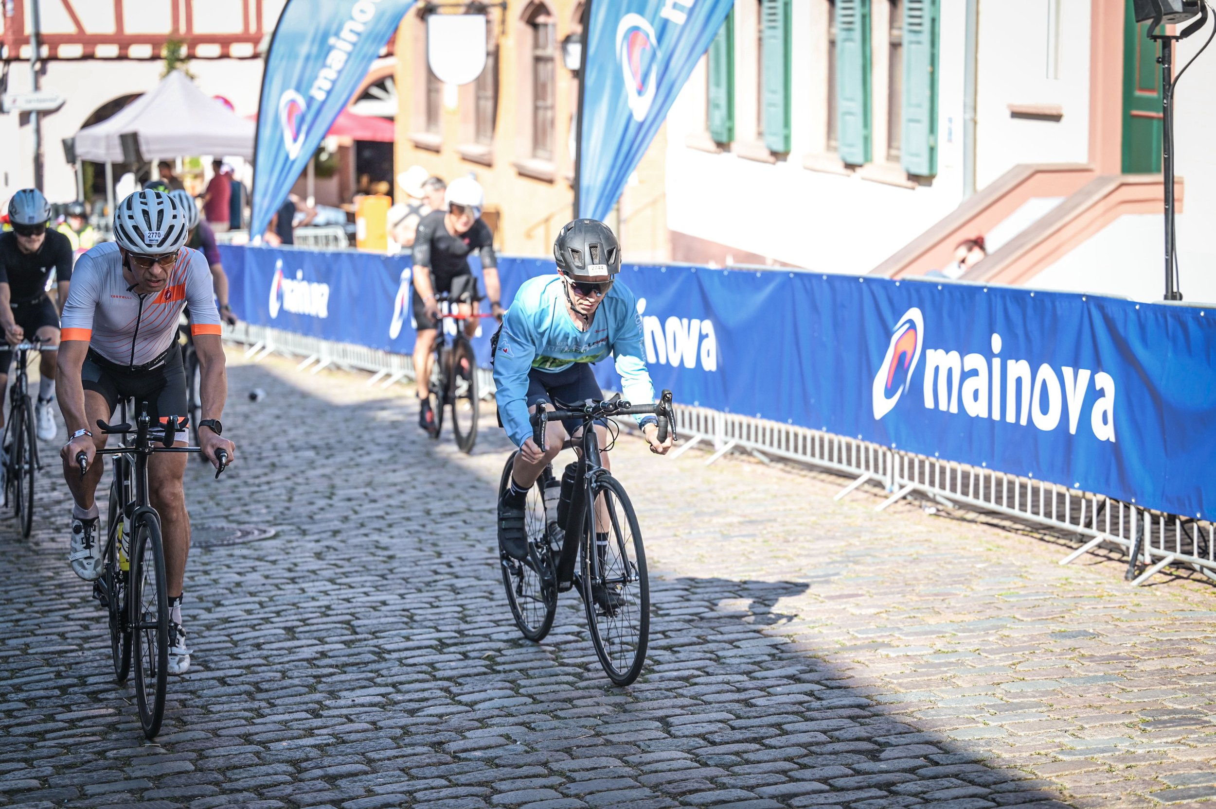 Mehrere Radfahrer, darunter ein älterer Mann, fahren auf einer Kopfsteinmahlstraße. Im Hintergrund sind blaue Banner mit dem Schriftzug 'Mainova' und einigen Gebäuden mit grünen Fensterläden sichtbar.