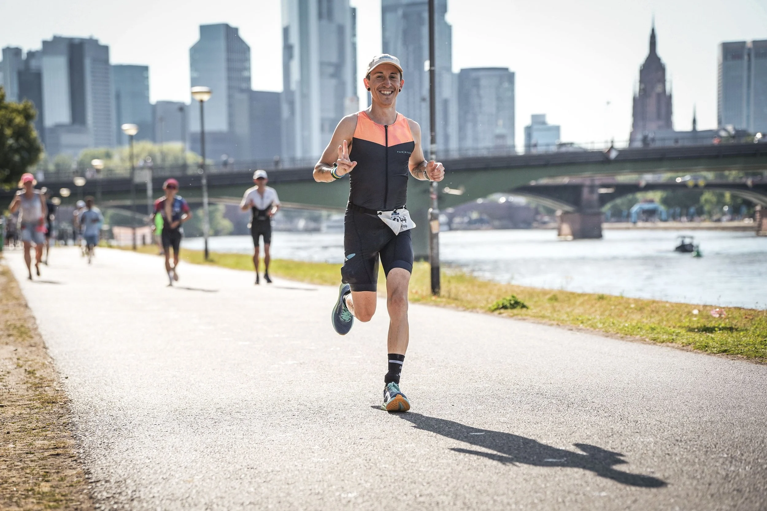 Mann joggt lachend an anderen Läufern entlang am Fluss in einer Stadt mit Hochhäusern im Hintergrund.