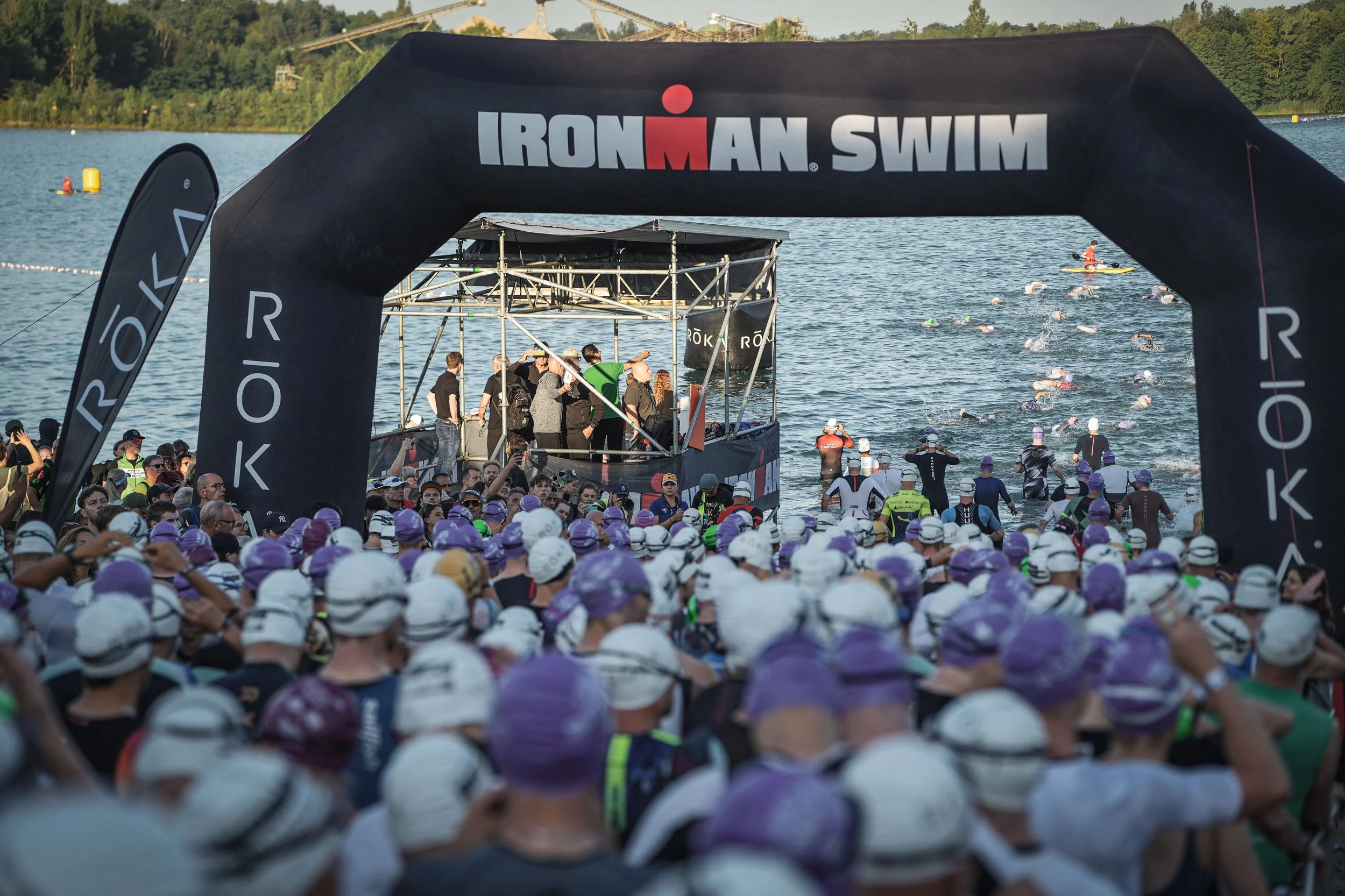 Start des Ironman-Wettbewerbs am Wasser mit zahlreichen Schwimmern, einer roten Ironman-Schwimm-Arch und Zuschauern am Ufer.
