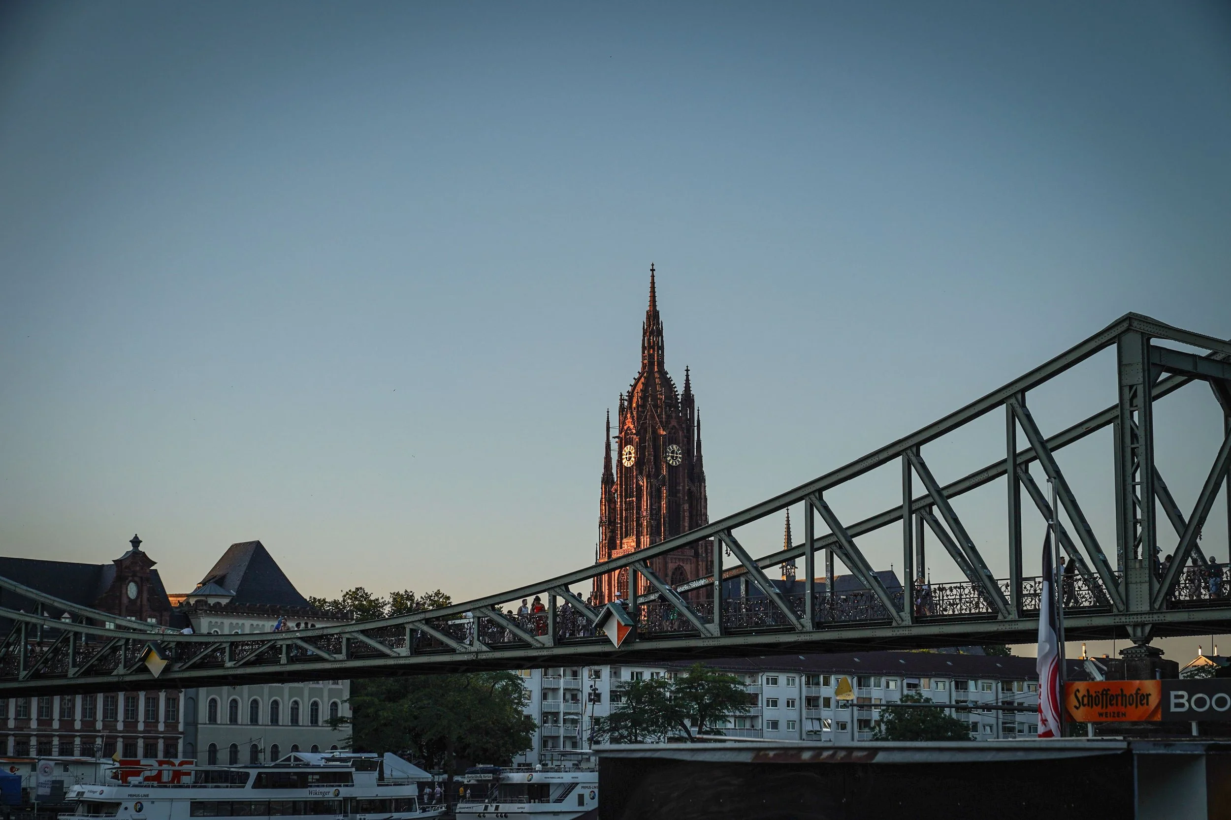 Blick auf eine Turmkirche bei Sonnenuntergang, mit einer gequerten Brücke im Vordergrund und einem Fluss mit Booten darunter.