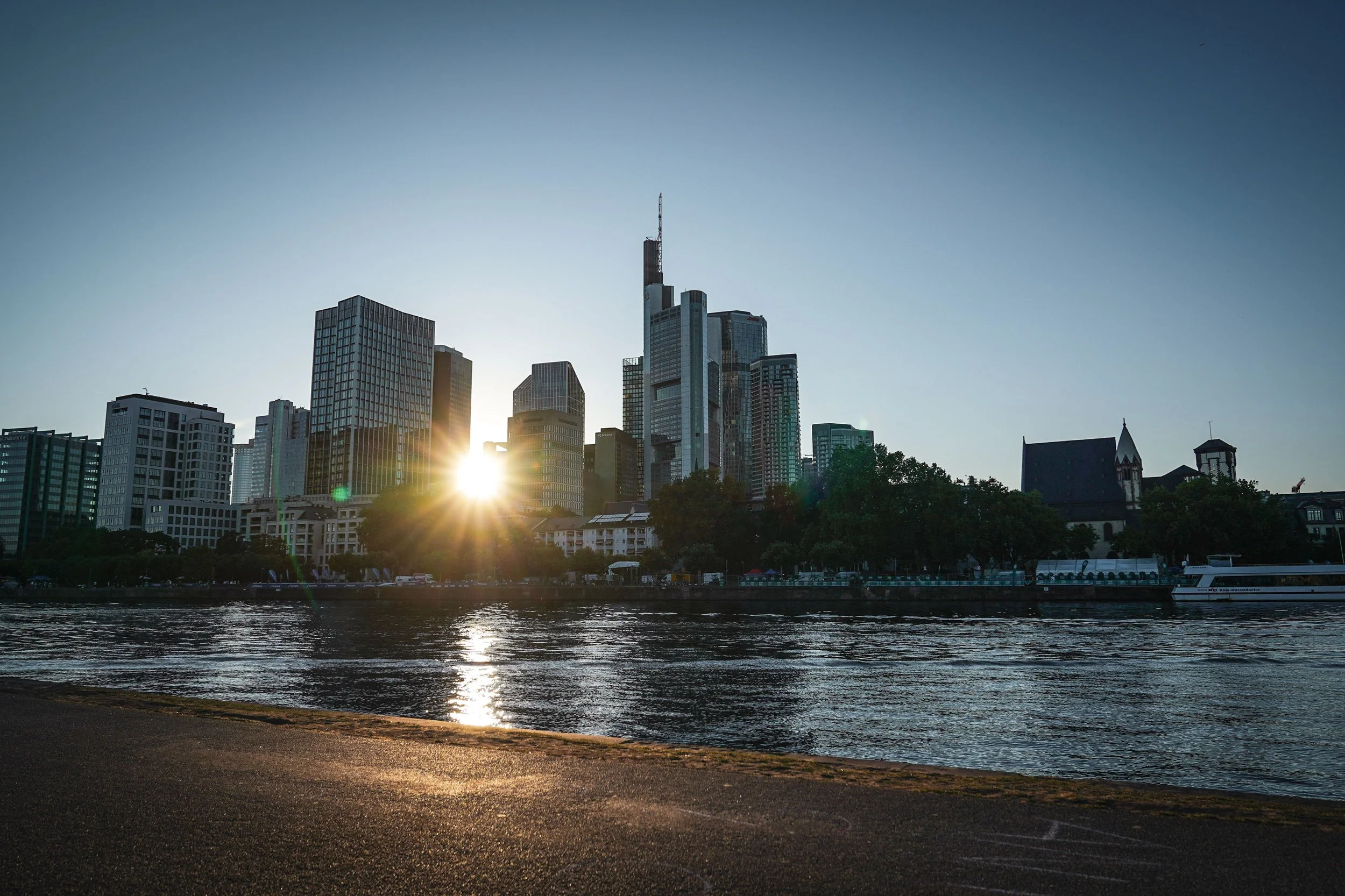 Stadt Skyline bei Sonnenuntergang über Fluss, mit modernen Hochhäusern und Kirche im Vordergrund.