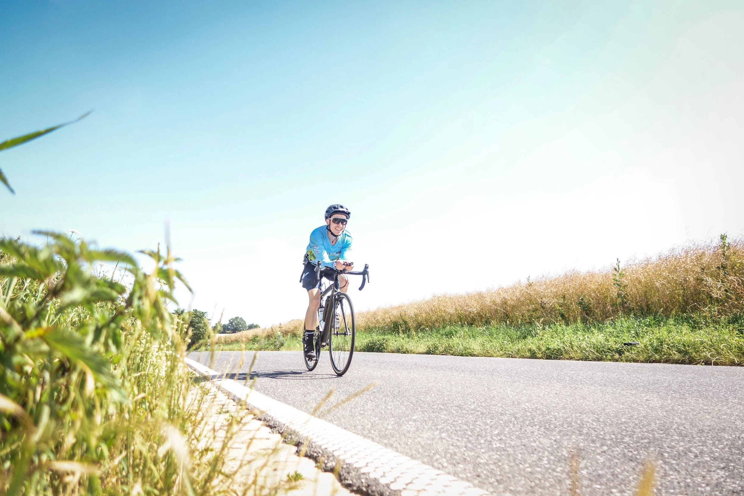 Ein Radfahrer in blauer Kleidung, Helm und Sonnenbrille fährt an einem sonnigen Tag auf einer Landstraße entlang, umgeben von Gräsern und Büschen.