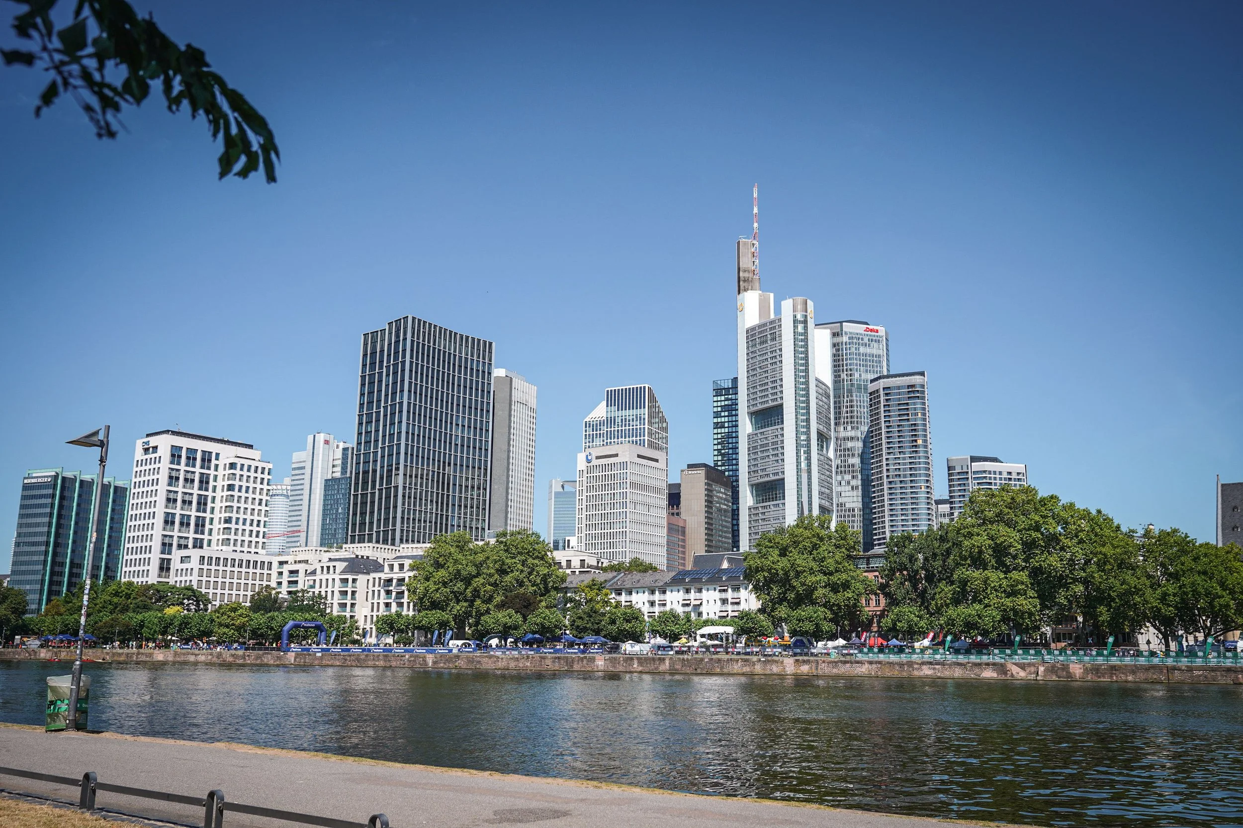 Die Aufnahme zeigt eine Skyline moderner Hochhäuser an einem sonnigen Tag, mit einigen Bäumen im Vordergrund und einer Wasserfläche im Vordergrund.