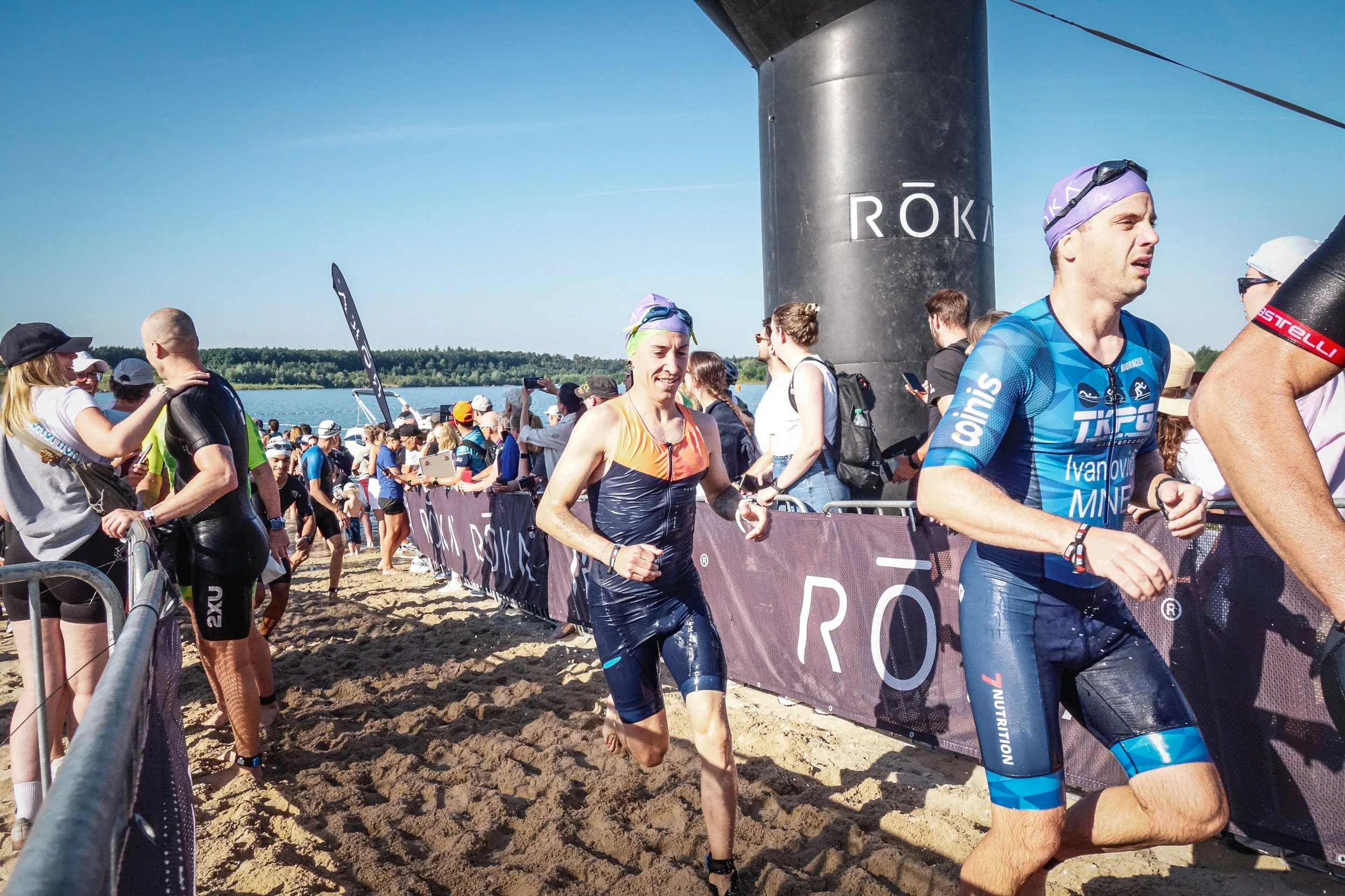 Marathonläufer bei einem Wettkampf am Ufer eines Sees, umgeben von vielen Zuschauern, bei sonnigem Wetter.