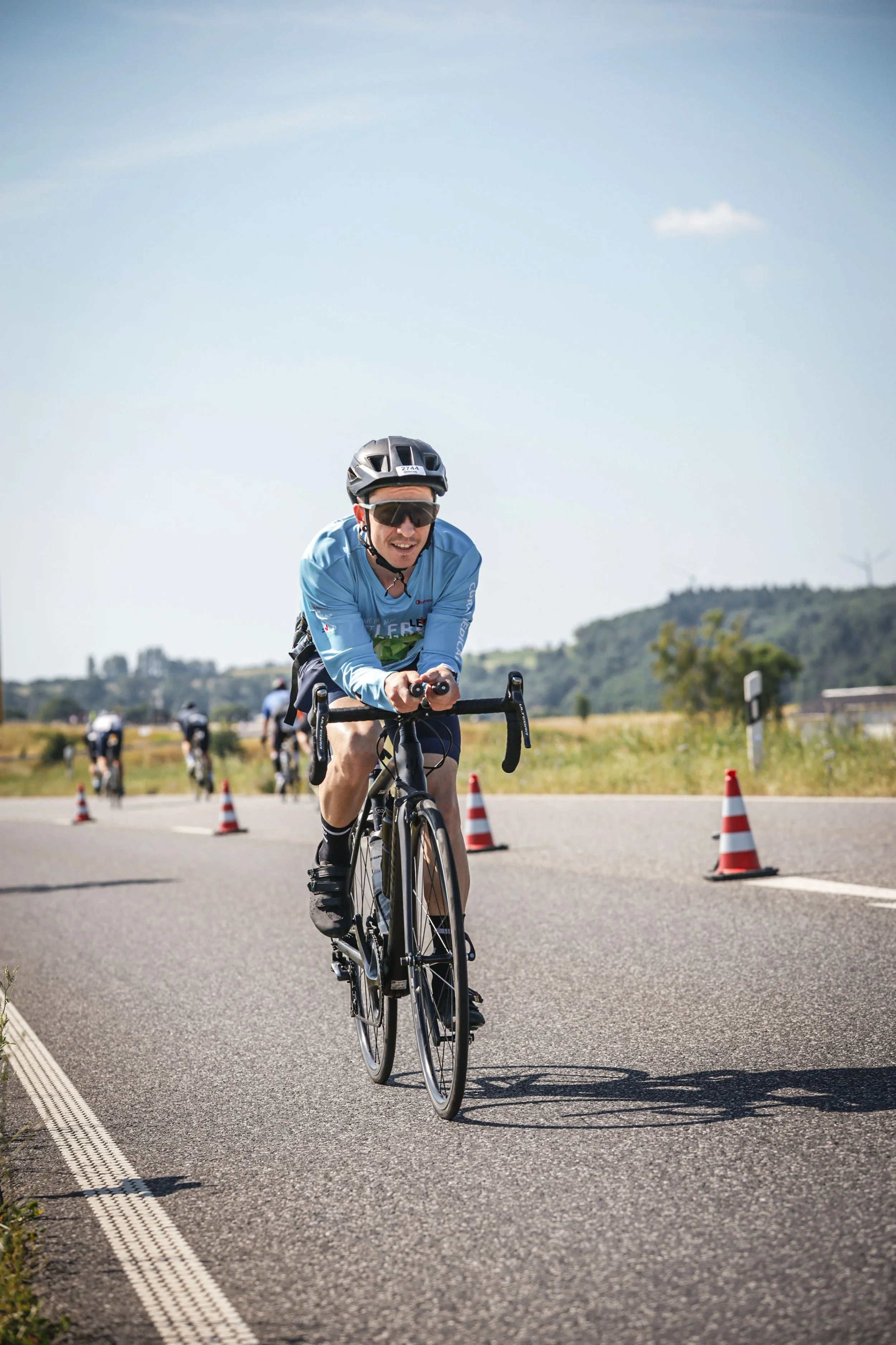 Mann fährt Fahrrad zwischen rote Verkehrsleitkegel auf einer Straße im Freien, trägt Helm und Sonnenbrille, im Hintergrund Hügel und Windkraftanlagen, sonniges Wetter.