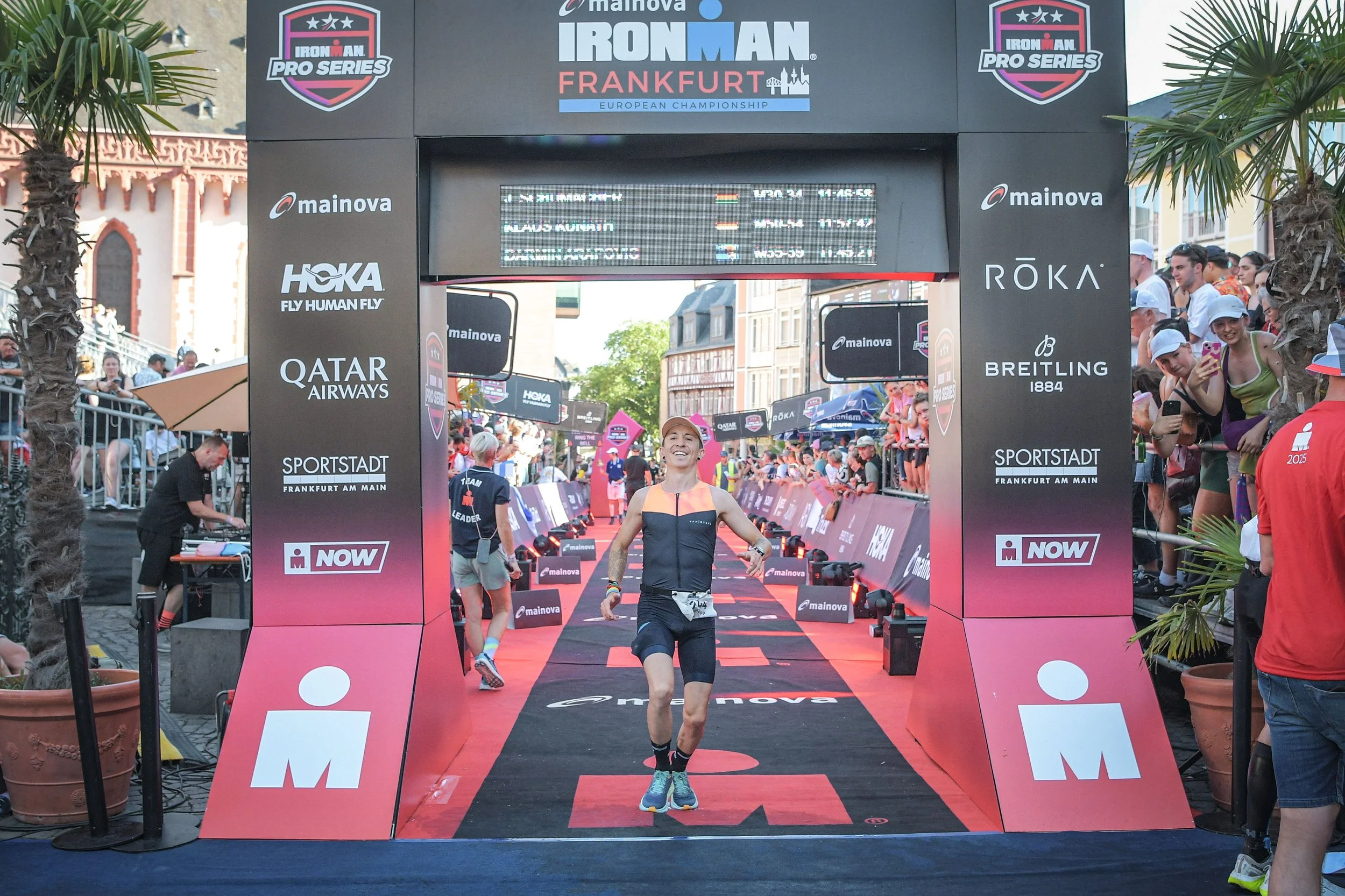 Ein Mann läuft bei einem Ironman-Wettbewerb durch die Ziellinie, umgeben von Zuschauern und Veranstaltungswerbung in Frankfurt am Main.
