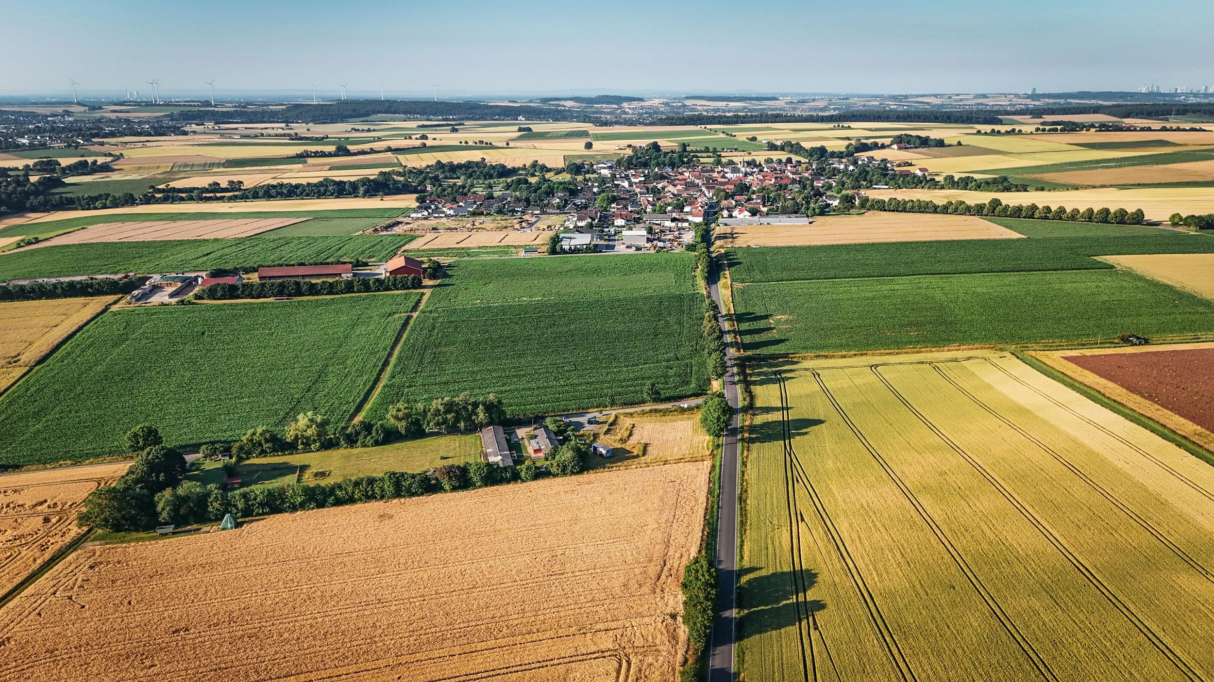Luftaufnahme einer ländlichen Landschaft mit Feldern, einem kleinen Dorf und Windkraftanlagen im Hintergrund.