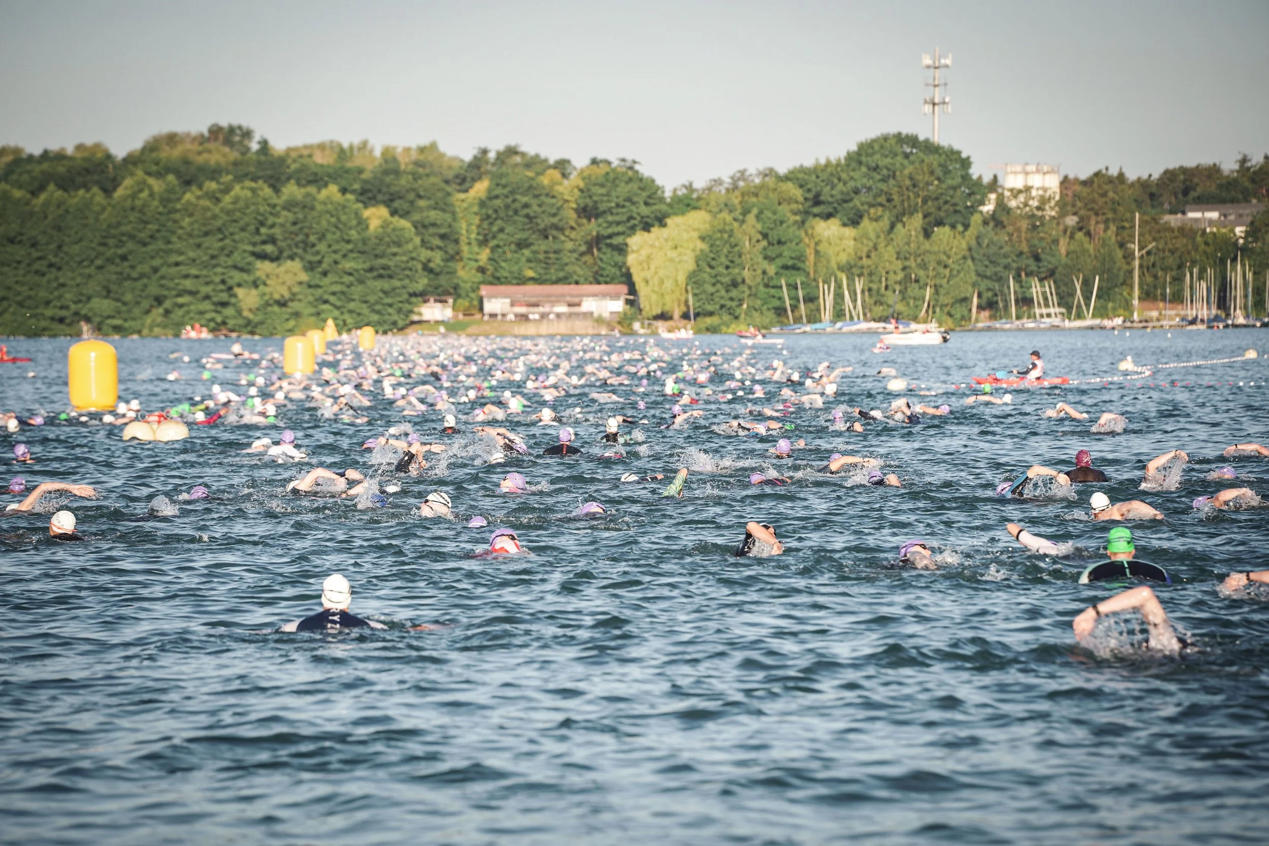 Viele Schwimmer bei einem offenen Wassertriathlon im See, mit Bäumen im Hintergrund und Booten am Ufer.