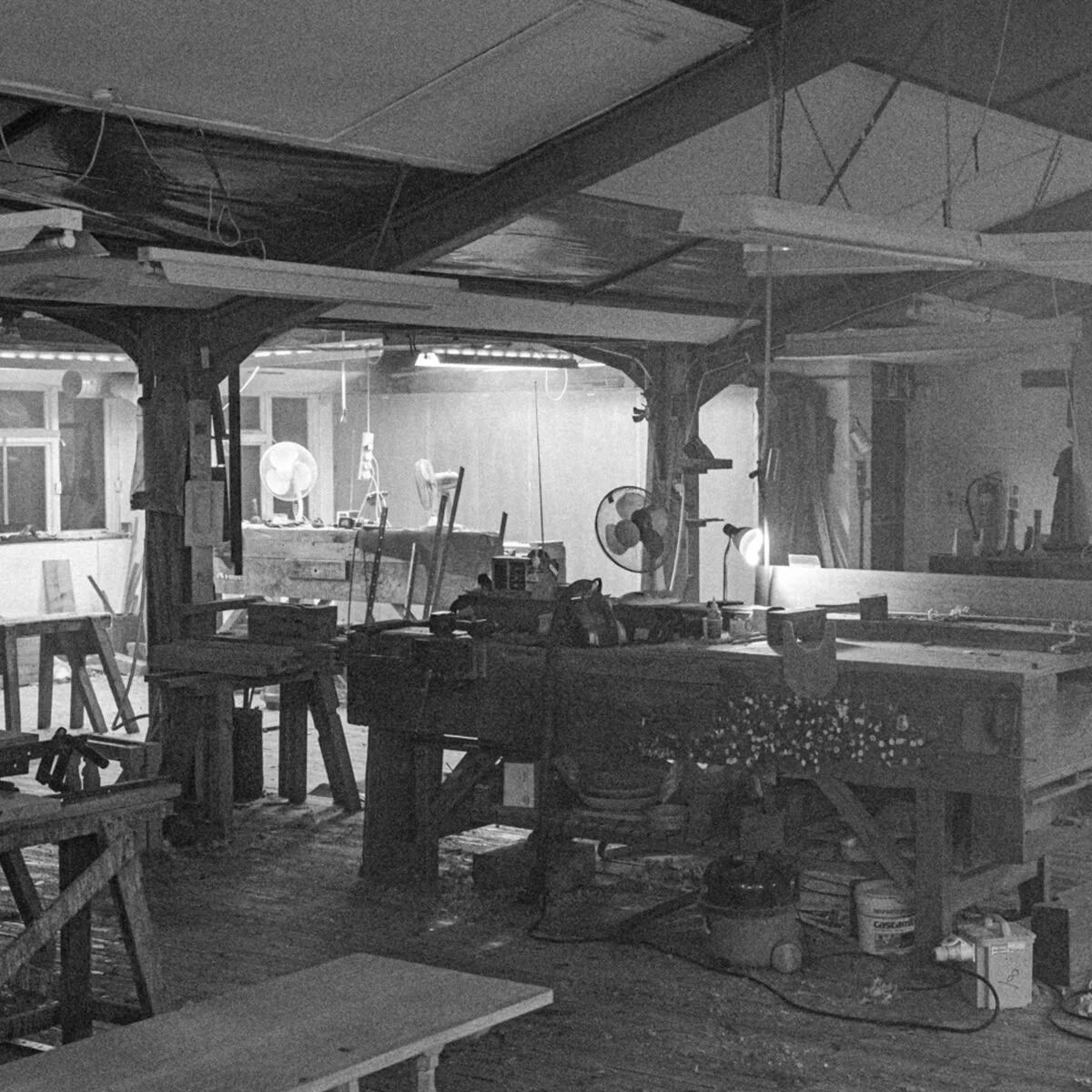 A cluttered woodworking shop with various tools, workbenches, and fans, seen in black and white.