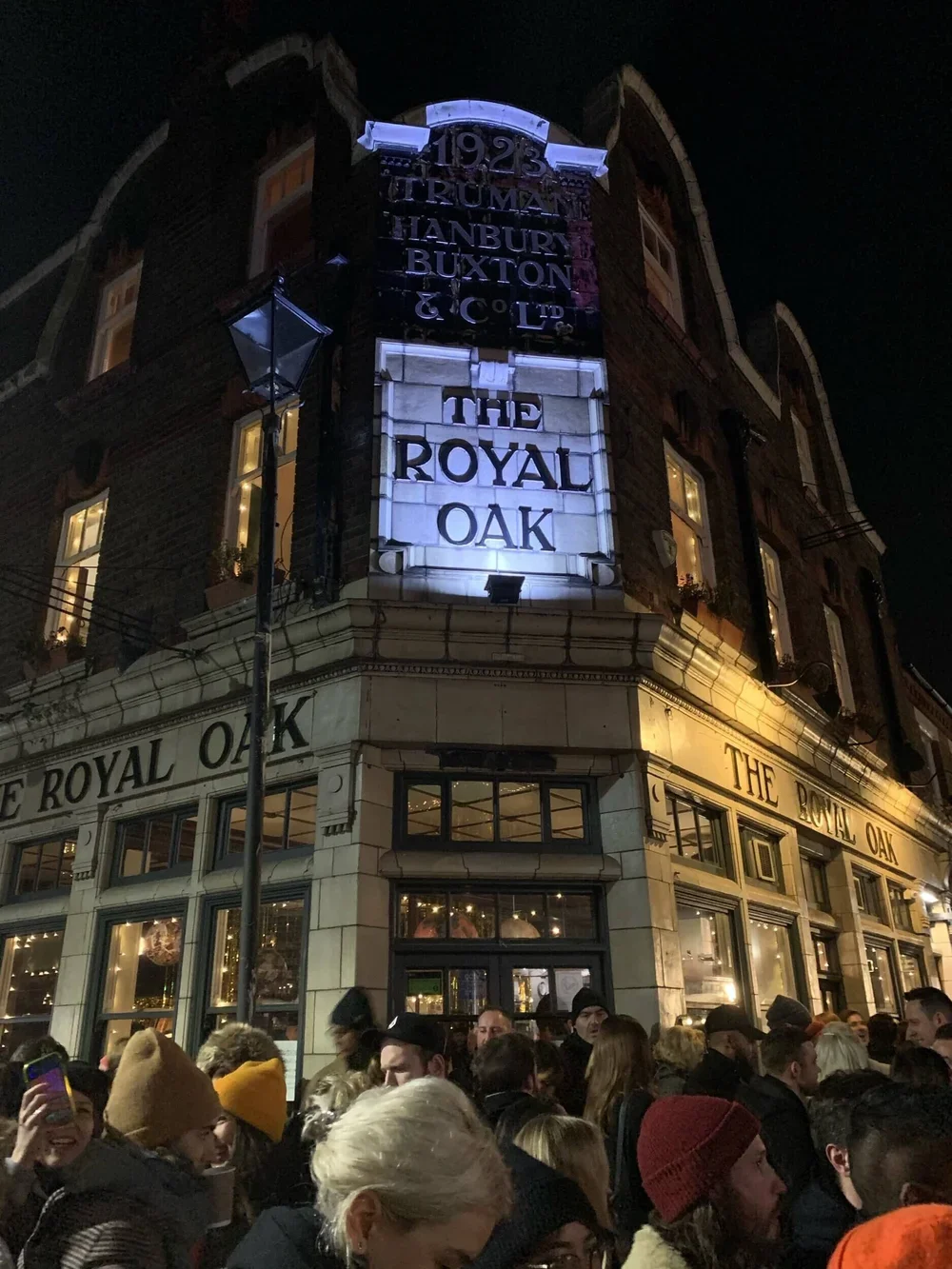 Nighttime scene outside The Royal Oak pub with a large crowd of people gathered in front. The building has brickwork, illuminated signs, and windows with interior lights. The signs indicate the pub's name and the year 1923.