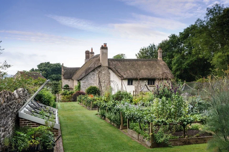 A picturesque cottage with a thatched roof surrounded by a lush garden with vegetable beds, trees, and a stone wall.