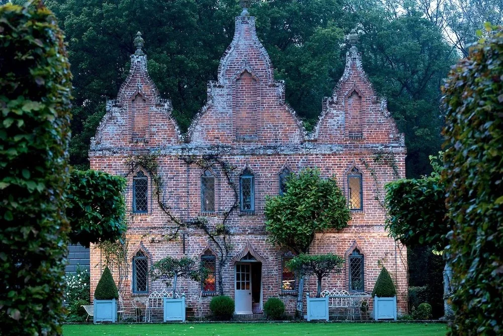A brick Gothic-style building with pointed arch windows, surrounded by trees and shrubs, with outdoor furniture in front.