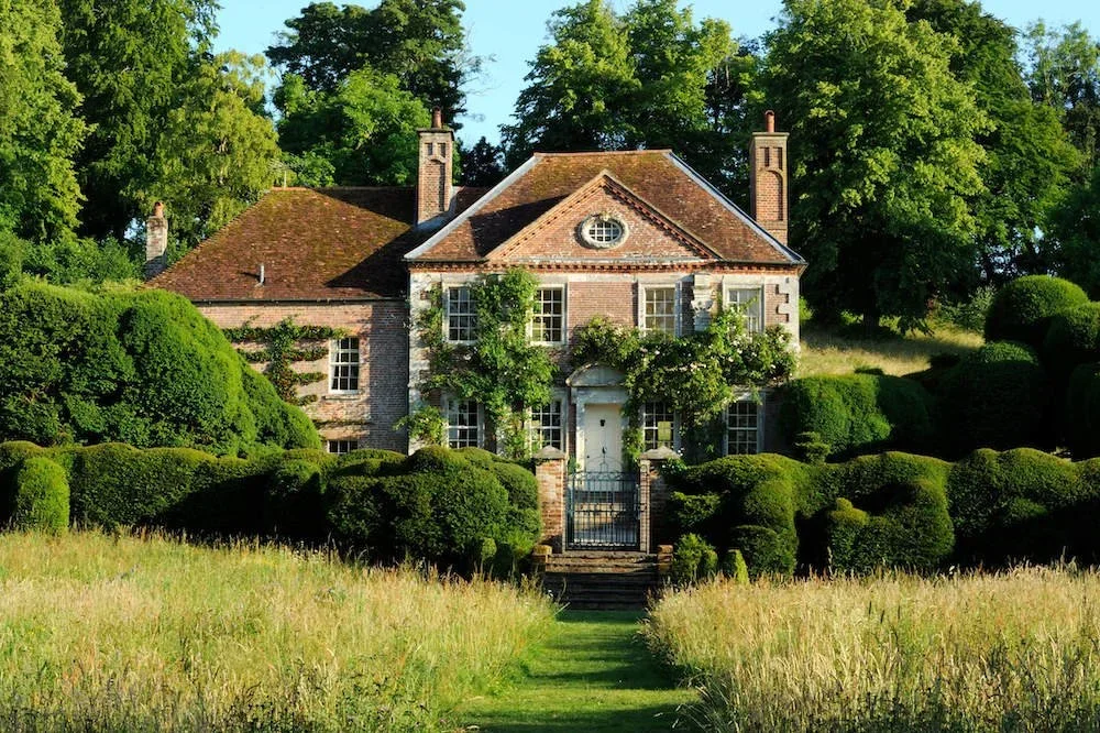 A large house surrounded by lush greenery, trees, and neatly trimmed bushes, with a pathway leading up to the front door.