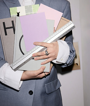 A person in a tailored grey suit holding a stack of colorful design samples and a metal ruler, suggesting creative planning, branding, or design work in a professional studio setting.