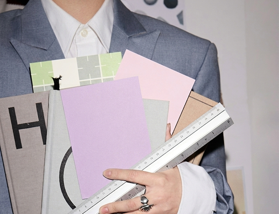 A person in a tailored grey suit holding a stack of colorful design samples and a metal ruler, suggesting creative planning, branding, or design work in a professional studio setting.