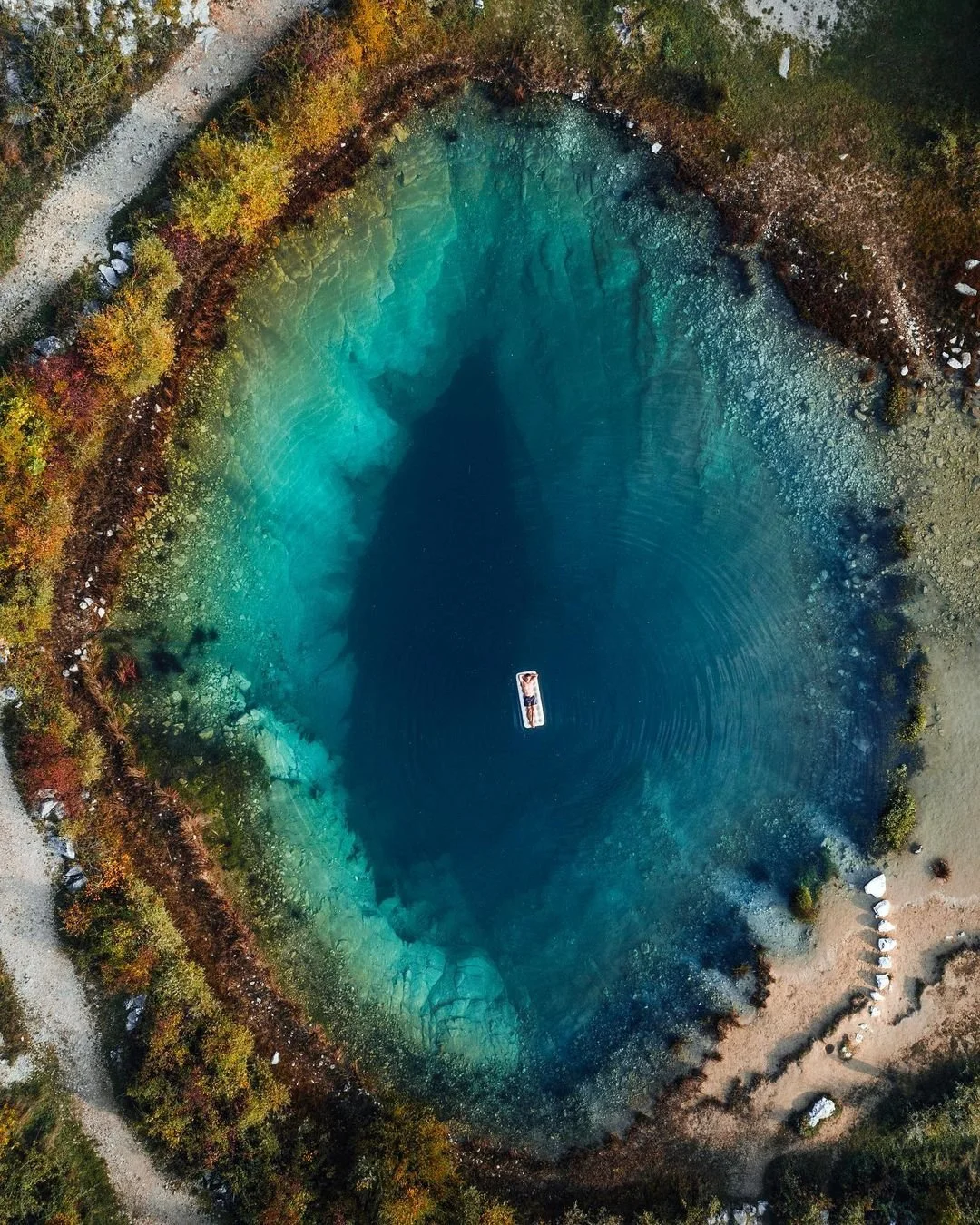 An aerial view of a vibrant blue, crater-like lake surrounded by colorful autumn trees and a dirt trail.