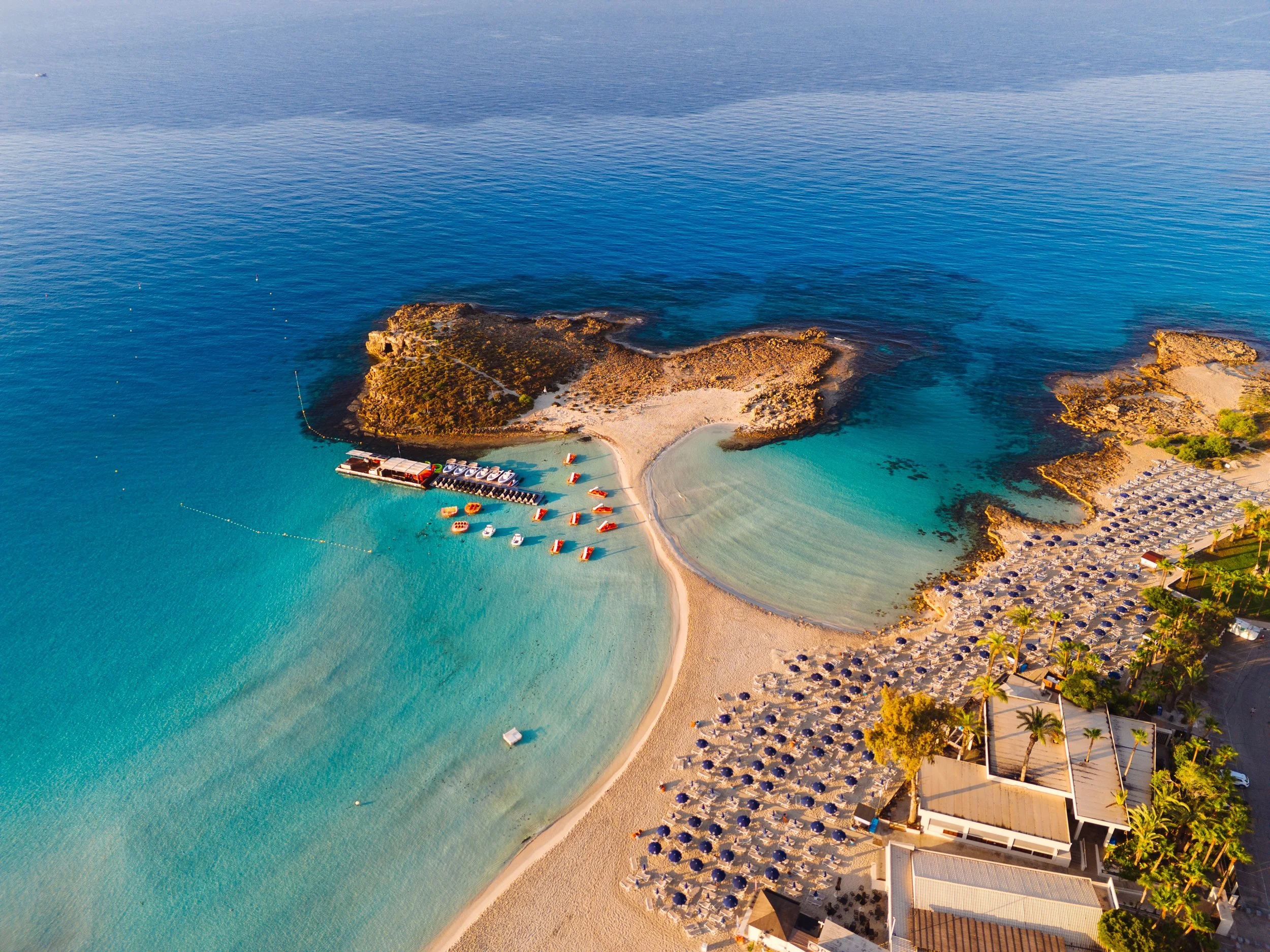 Aerial view of a tropical beach with turquoise water, sandy shore, small rocky islands, and beachfront buildings with umbrellas and palm trees.